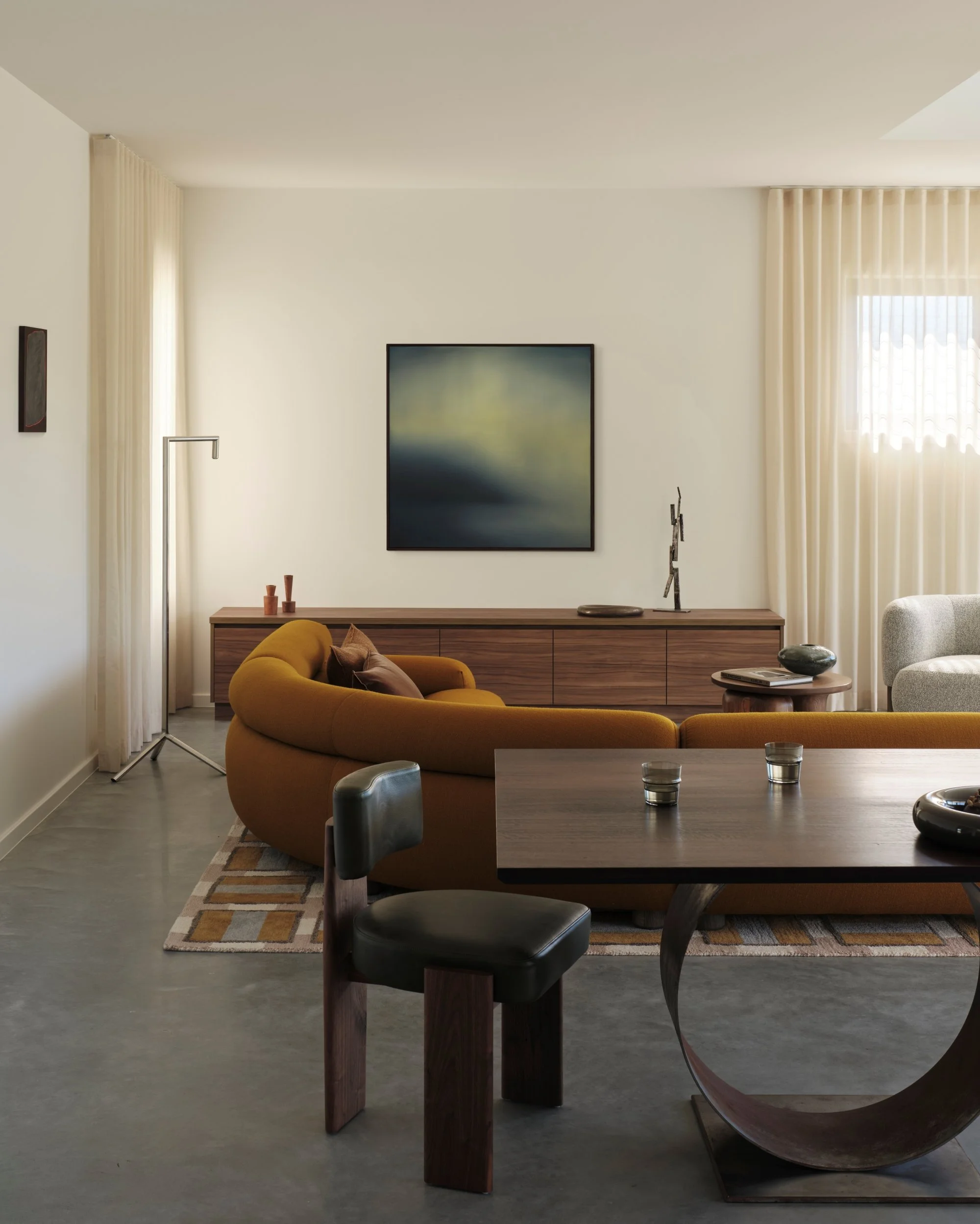 Open-plan living and dining space with sculptural dining table, curved sofa, timber cabinetry and contemporary artwork, highlighting warm textures and modern residential interior design.