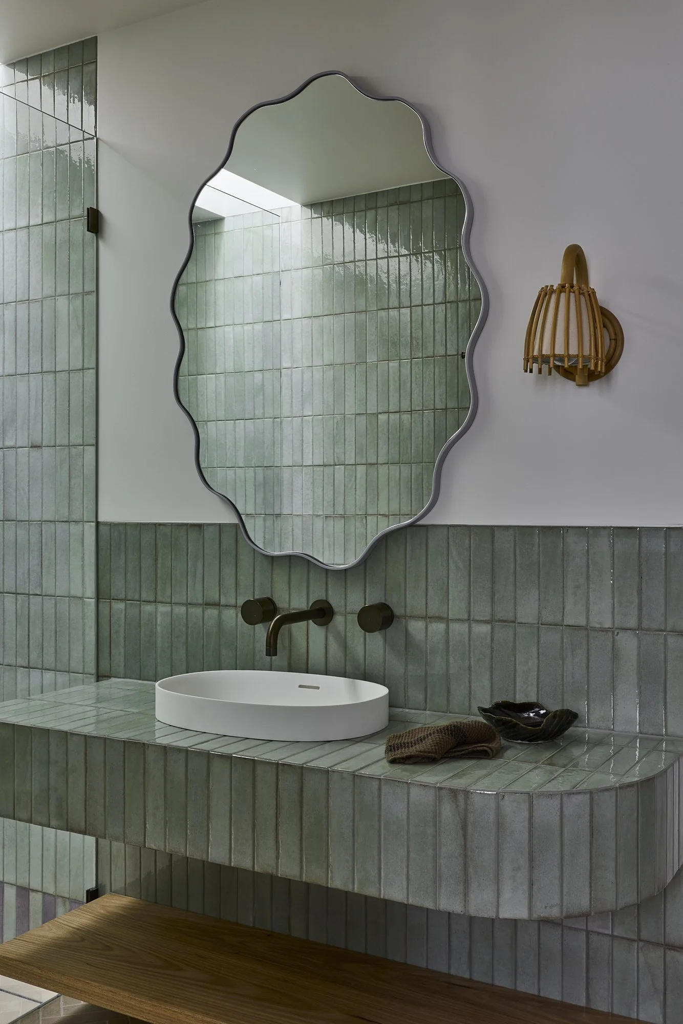 Green tiled bathroom vanity with sculptural curved mirror, white vessel basin, brushed brass wall-mounted tapware and handcrafted rattan wall light in a contemporary Australian coastal home.