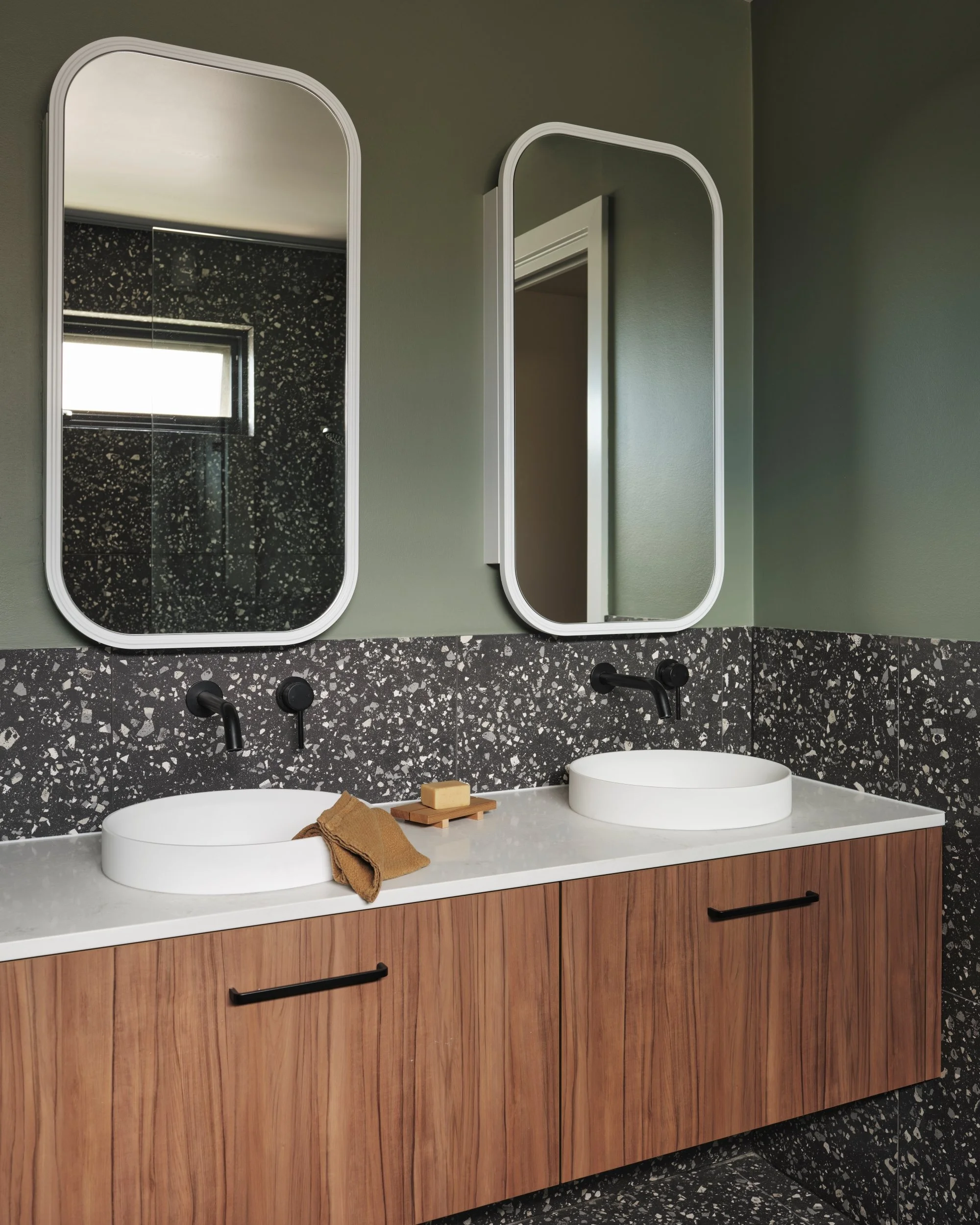 Modern bathroom with terrazzo wall tiles, olive green walls, twin round basins, wall-mounted black tapware and timber vanity cabinetry.