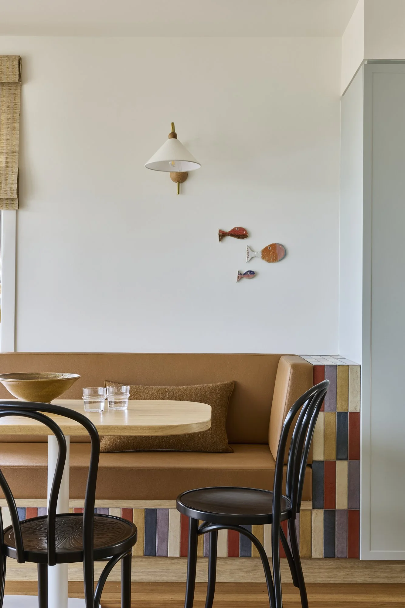 Built-in leather dining banquette with handcrafted colourful tiled detailing, timber dining table and decorative wall lighting in a warm contemporary coastal interior.