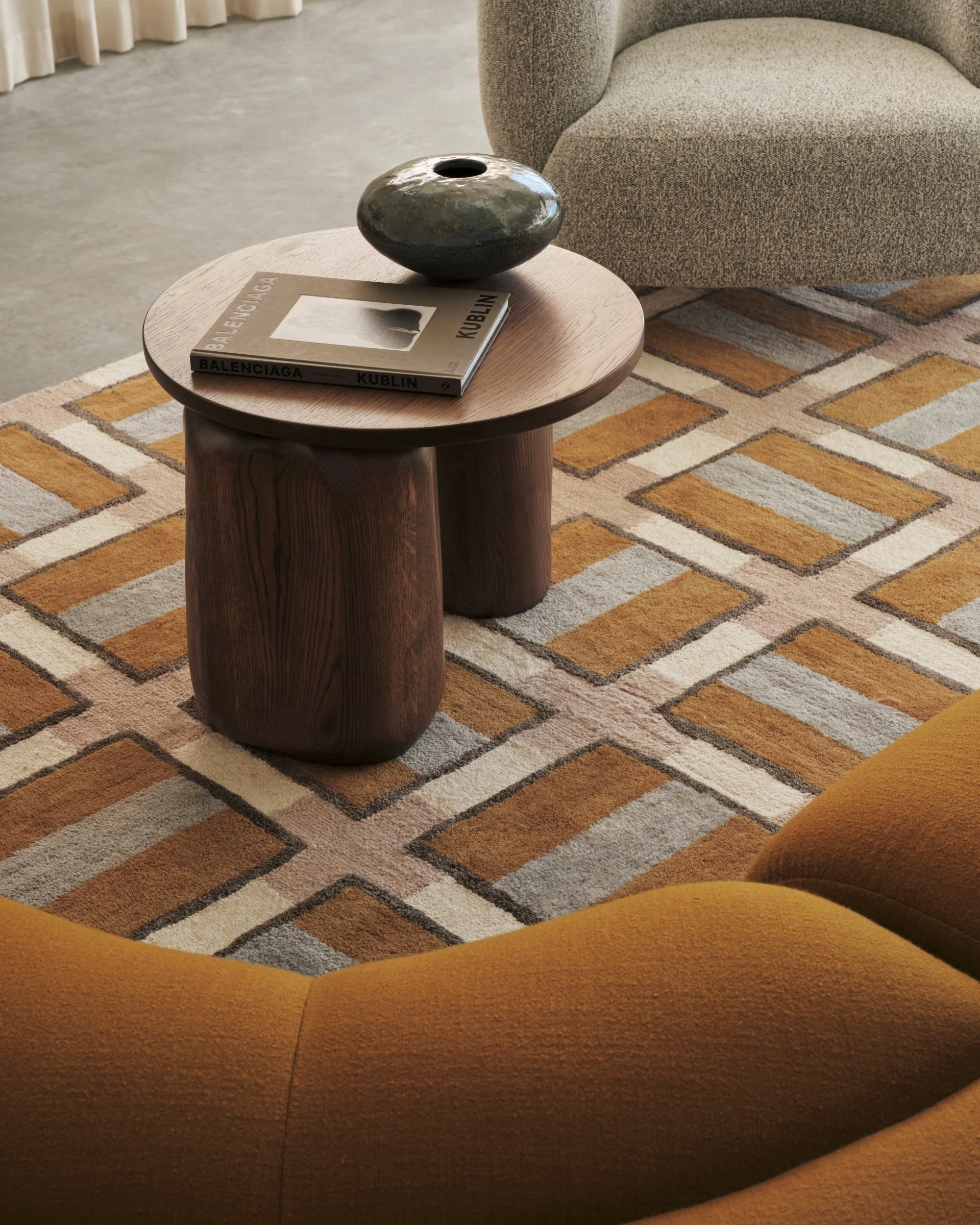 Timber coffee table with ceramic vase and design book set on a geometric patterned rug beside a textured armchair, highlighting modern furniture and warm interior styling.