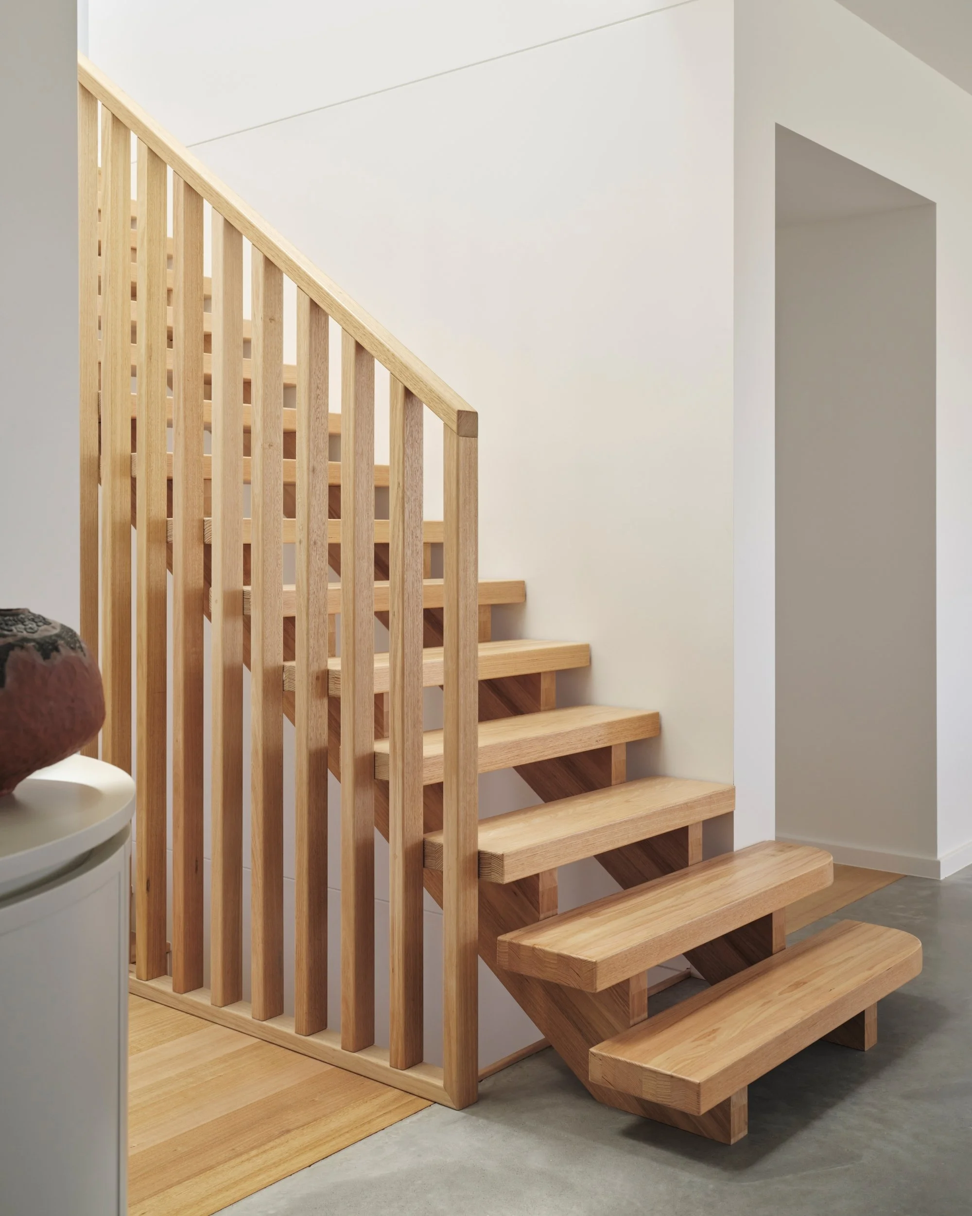 Minimalist timber staircase with solid oak treads and vertical balustrade set against white walls and polished concrete flooring in a contemporary interior.