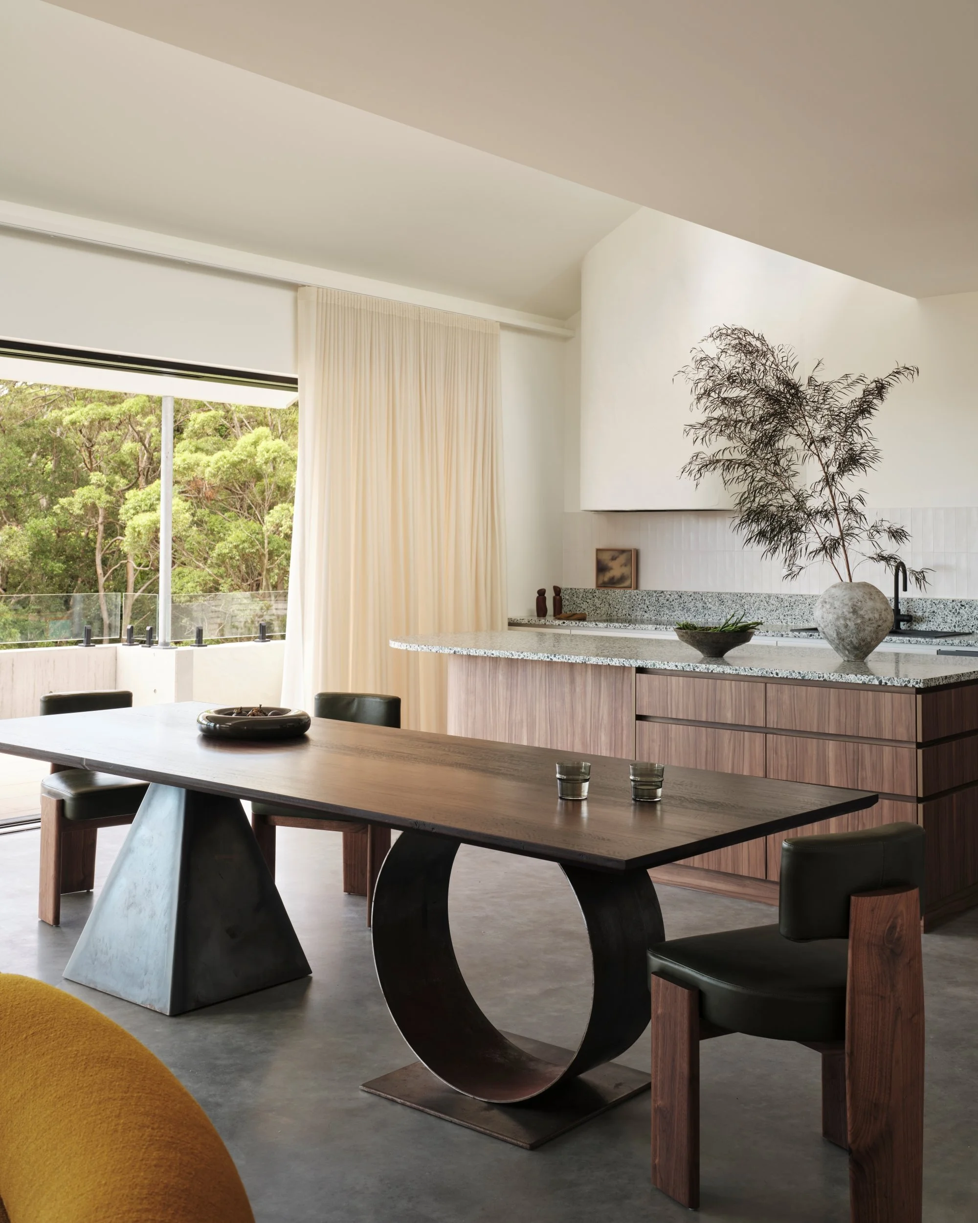 Open-plan dining and kitchen area with sculptural dining table, timber chairs, terrazzo island bench and soft sheer curtains overlooking greenery, capturing modern coastal interior architecture.