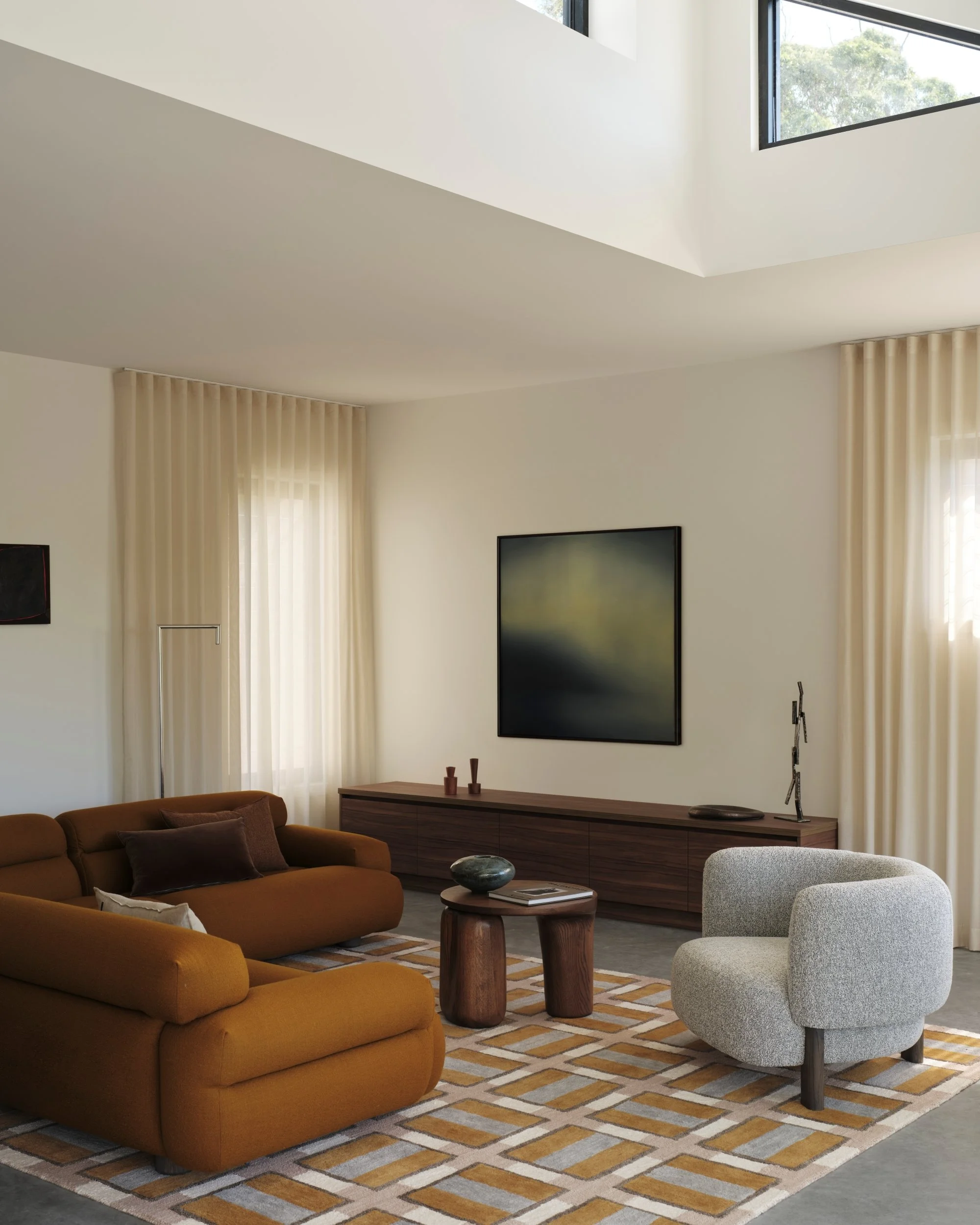 Modern living room with burnt orange sofa, textured armchair, patterned rug and timber sideboard beneath contemporary artwork, illuminated by soft daylight through sheer curtains.