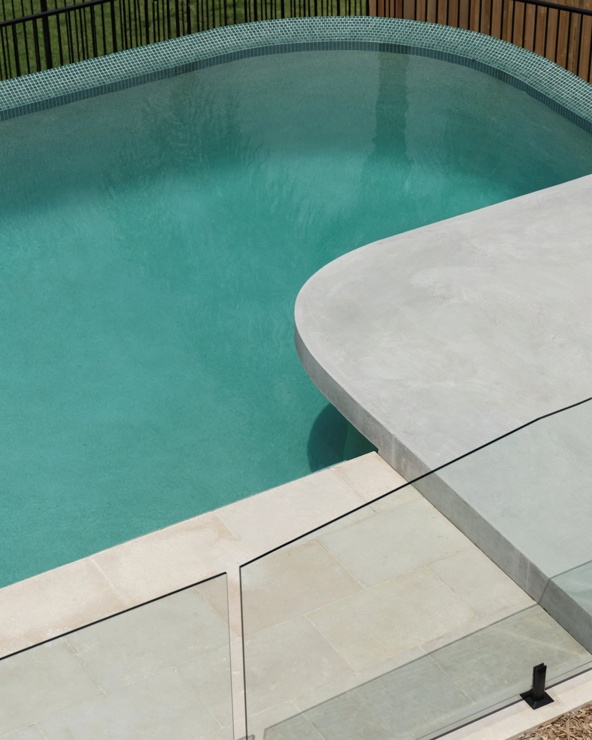 Curved concrete pool edge with pale stone paving and glass pool fence overlooking turquoise water, highlighting sculptural outdoor design.