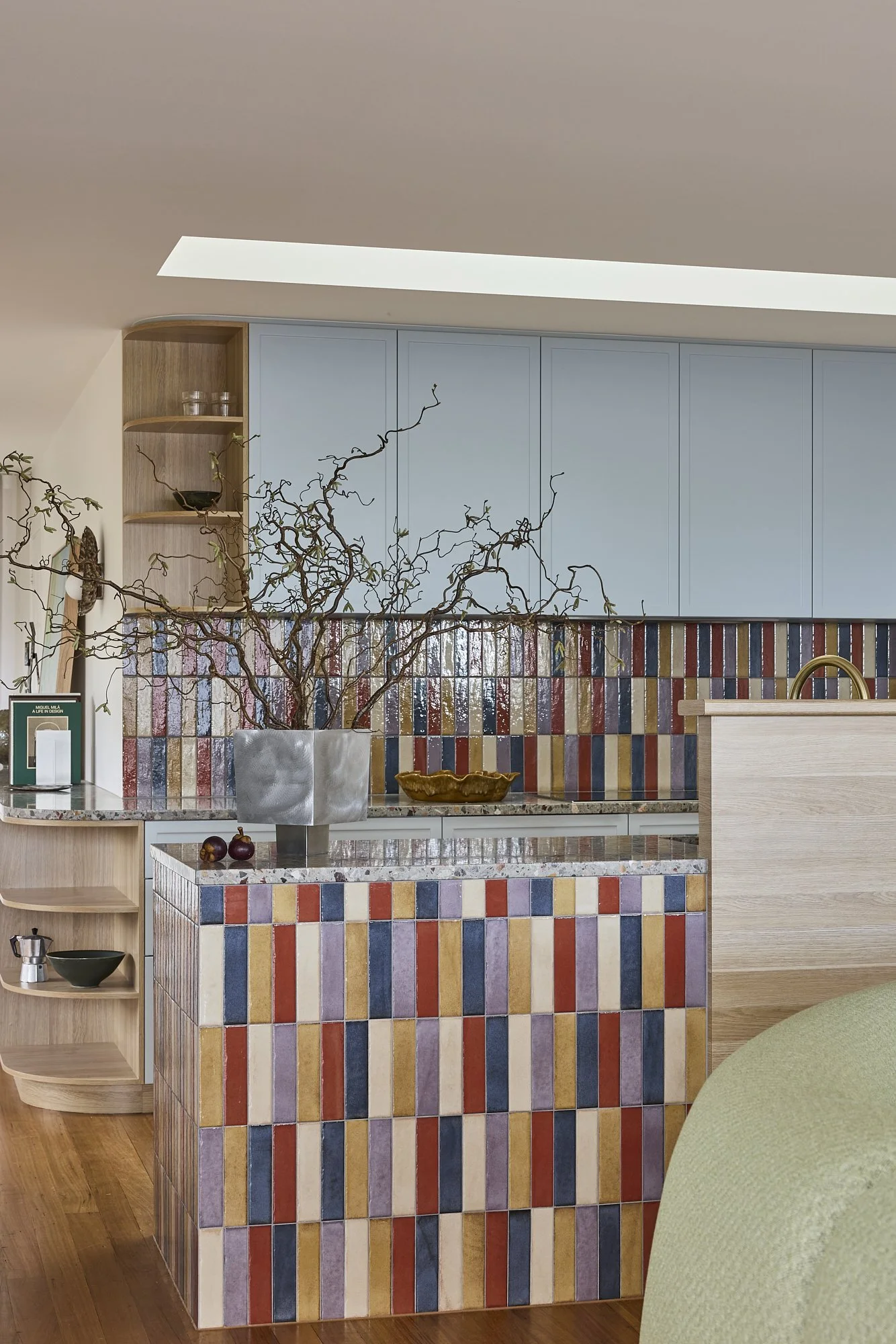 Contemporary kitchen featuring colourful tiled splashback, terrazzo benchtops, pale blue cabinetry and integrated timber shelving in a modern Australian coastal home.