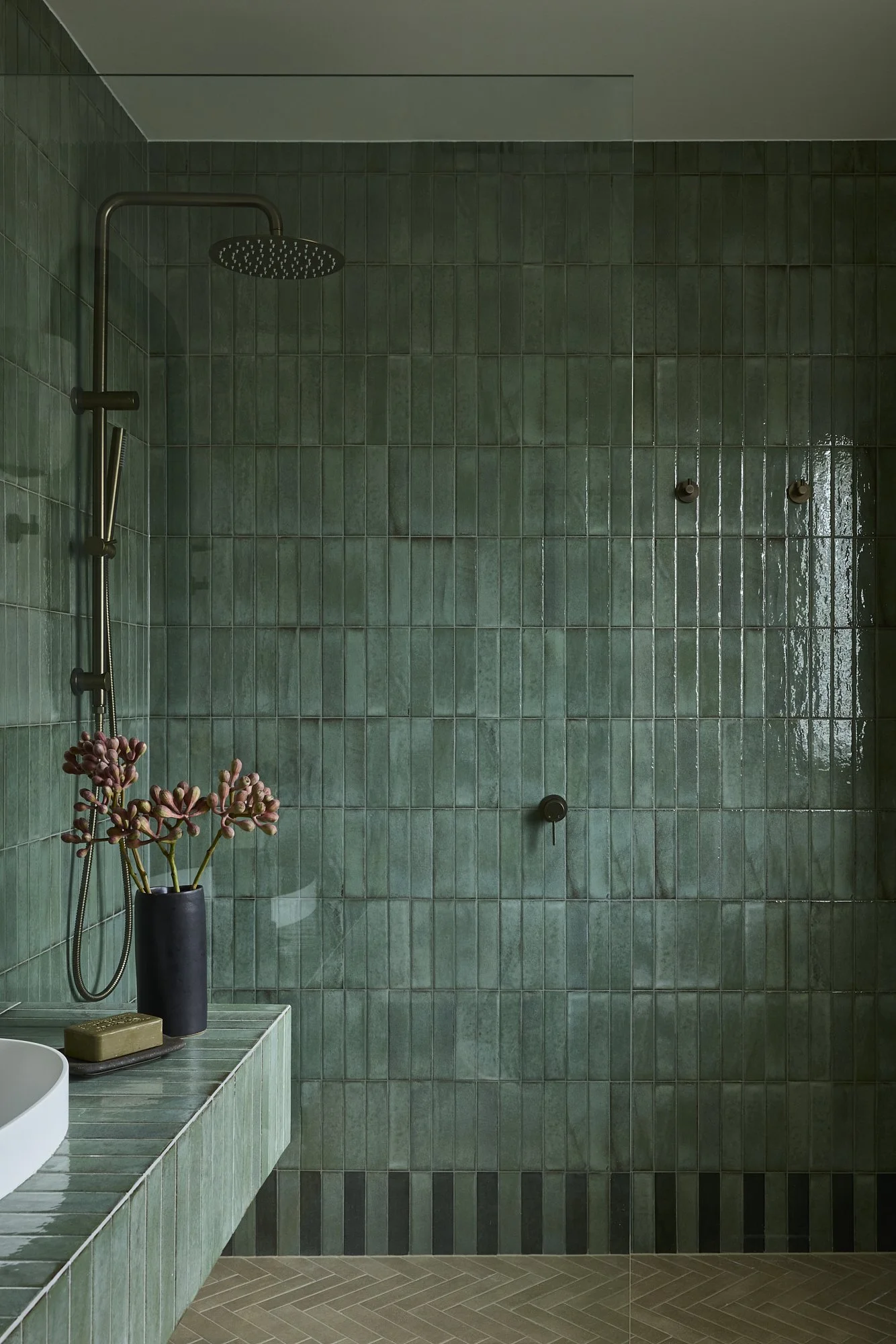 Walk-in shower with handcrafted green ceramic wall tiles, brushed brass rain shower, frameless glass screen and herringbone floor tiles in a contemporary Australian bathroom.
