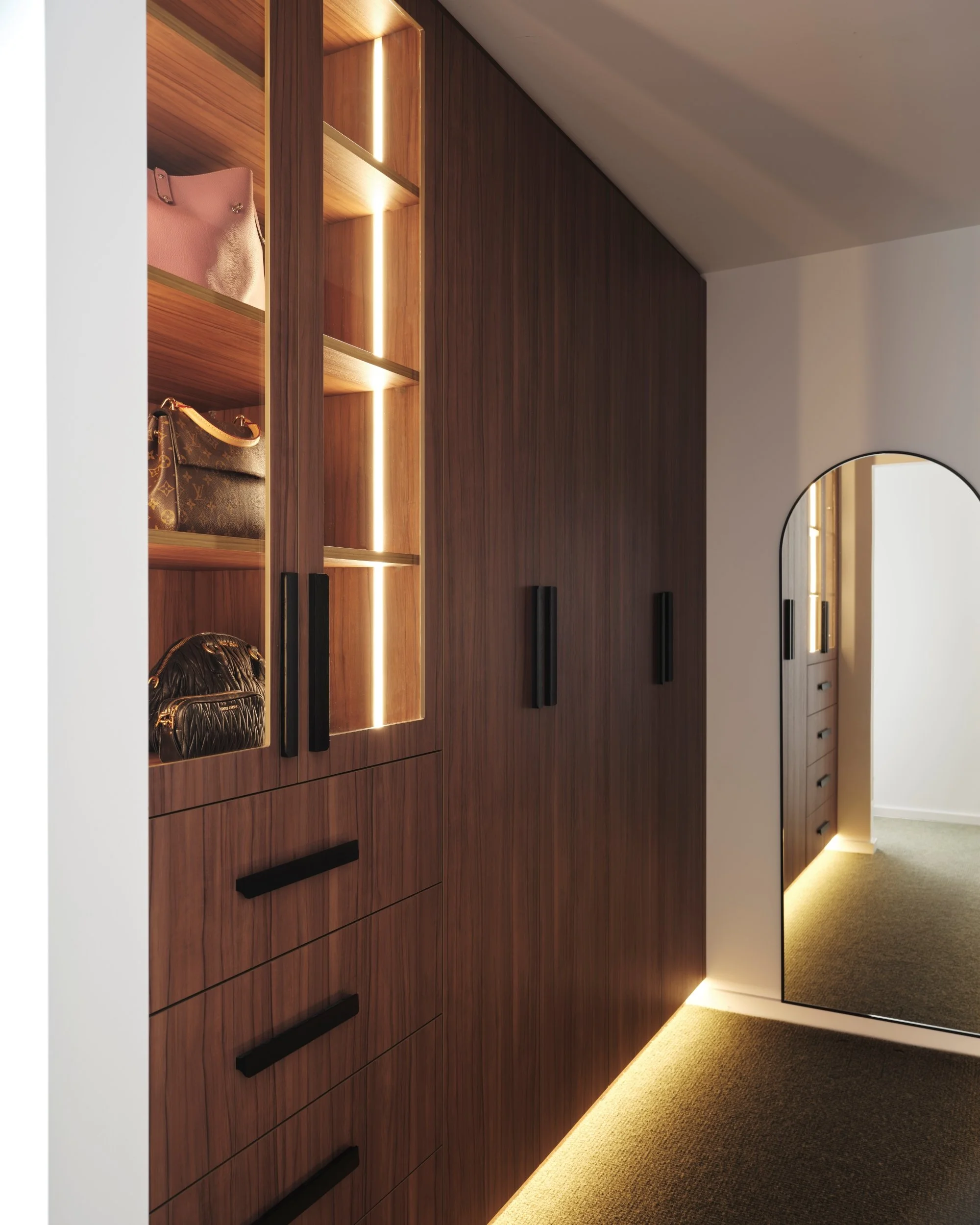 Custom walk-in robe with walnut cabinetry, illuminated display shelves and full-height mirror, showcasing bespoke storage and integrated lighting.