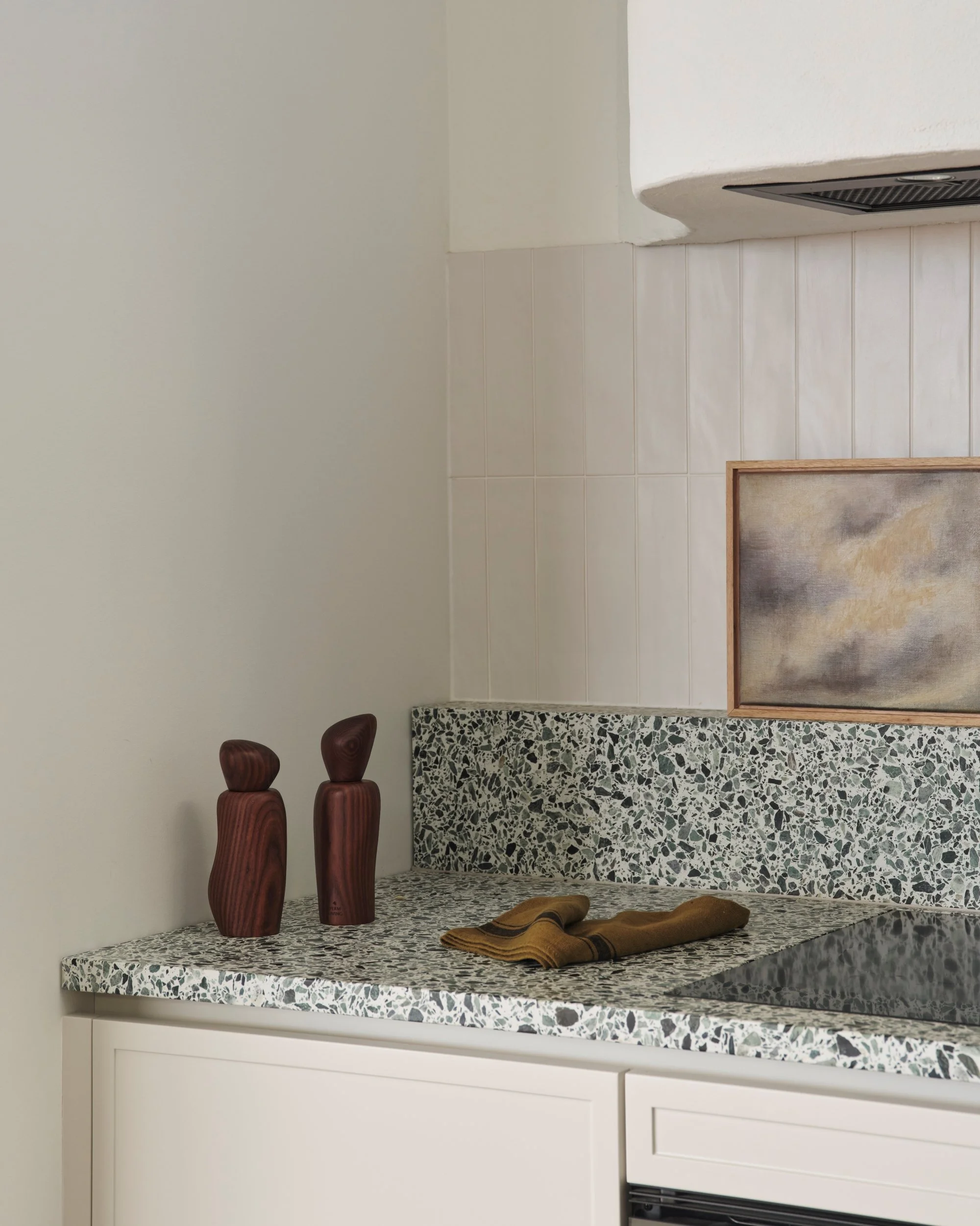 Minimalist kitchen bench with terrazzo splashback, timber accessories and framed artwork beneath a sculptural plaster range hood in a contemporary interior kitchen design.