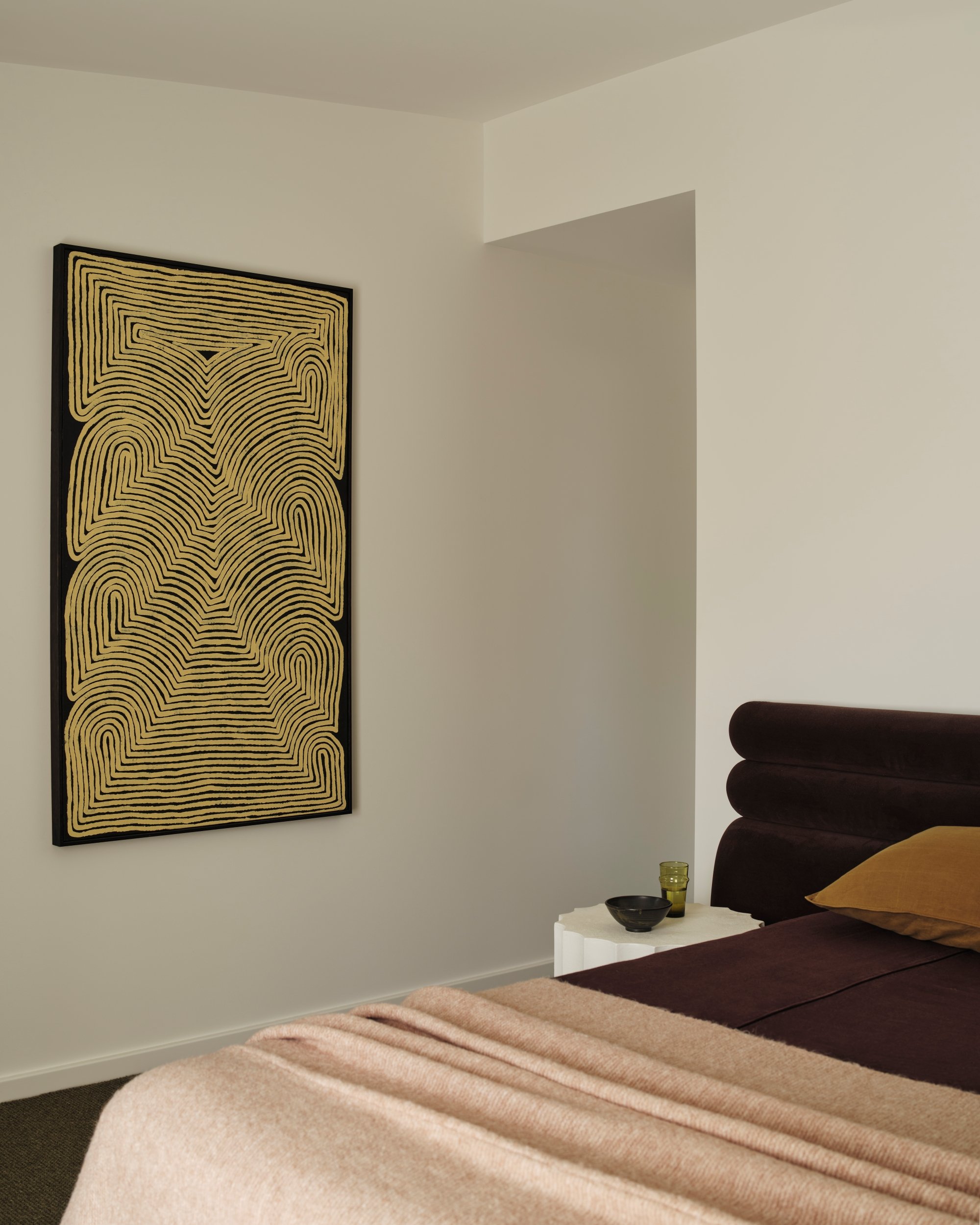 Bedroom corner featuring leather bedhead, sculptural bedside table and textured wall artwork in warm neutral tones.
