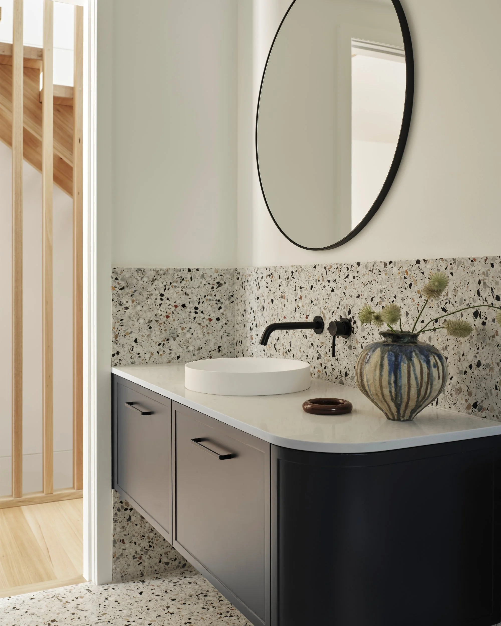 Compact powder room with terrazzo splashback, round basin, black wall-mounted tapware and curved vanity joinery beneath a circular mirror.
