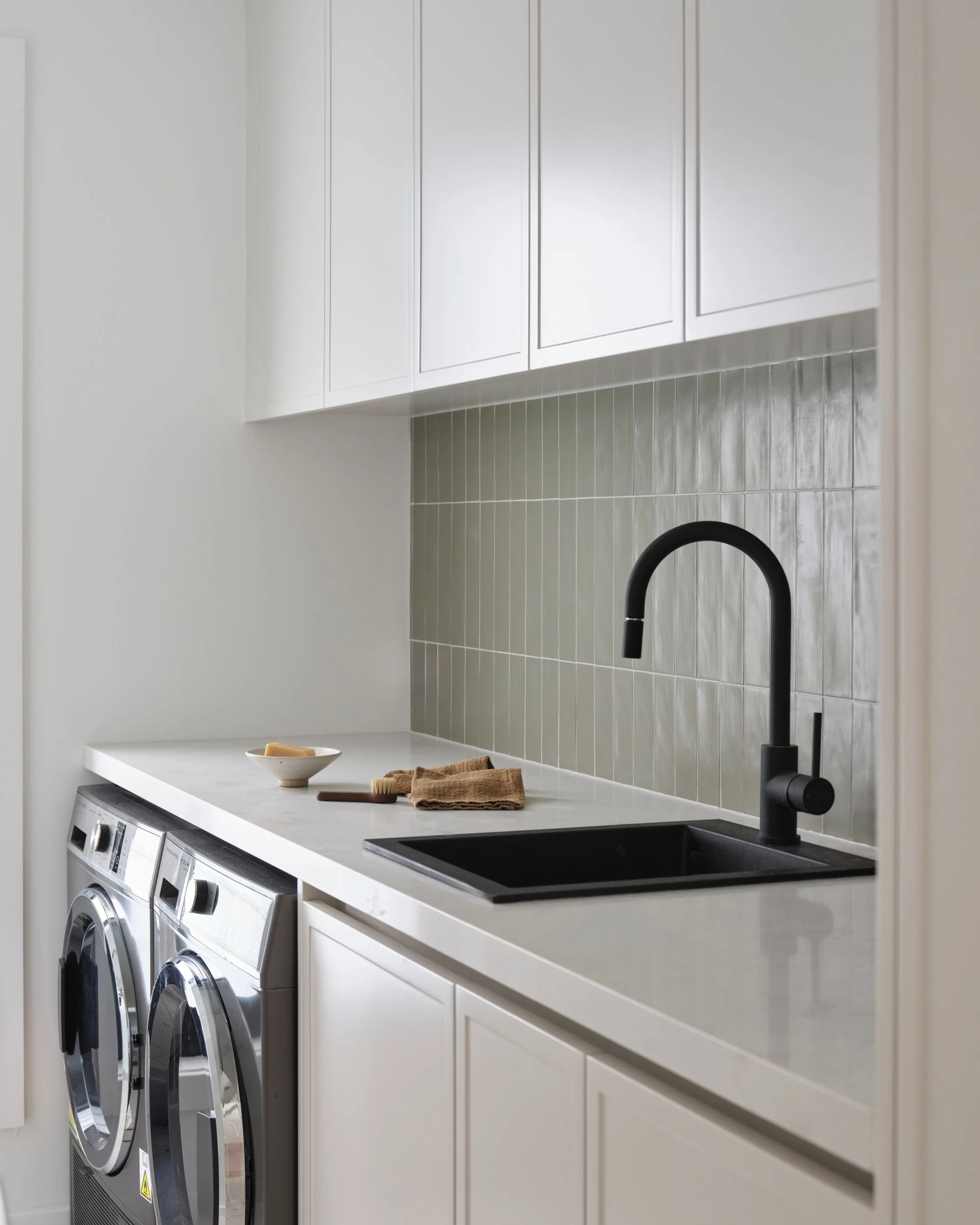 Modern laundry with white cabinetry, green tiled splashback, matte black tapware and integrated washing machines beneath a stone benchtop, showcasing clean contemporary utility room design.