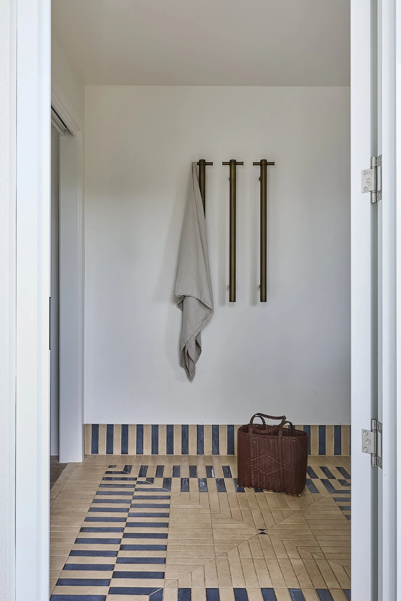 Modern coastal laundry with patterned beige and navy floor tiles, matching striped skirting tiles, aged brass heated towel rails, white walls and woven basket in a minimalist Australian home by Revel Design.