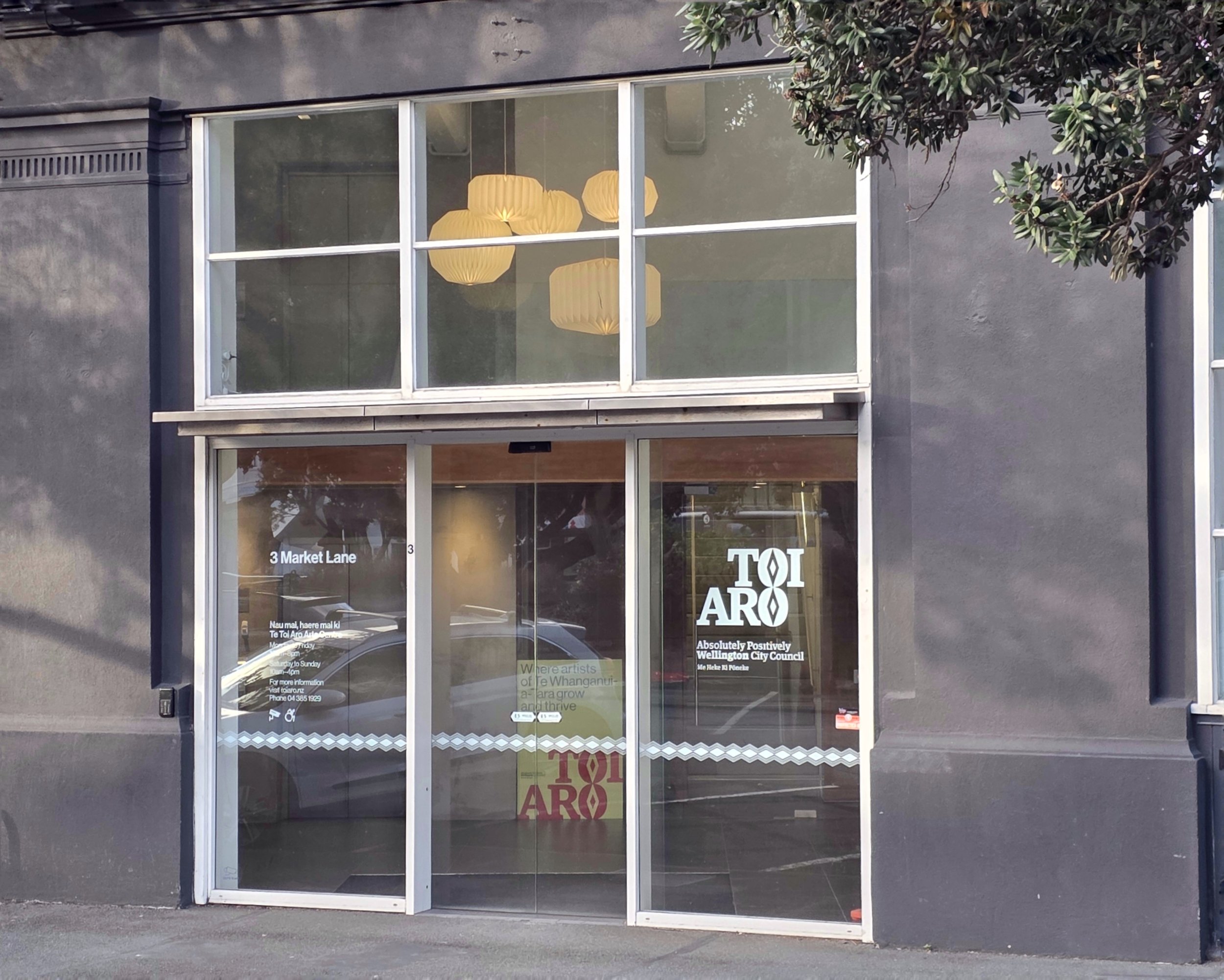 View of a modern storefront with large glass windows and door, with reflections of trees and street outside. Inside, hanging yellow pendant lights are visible. Signs on the door indicate location at 3 Market Lane, Wellington City Council, and promotional messages.