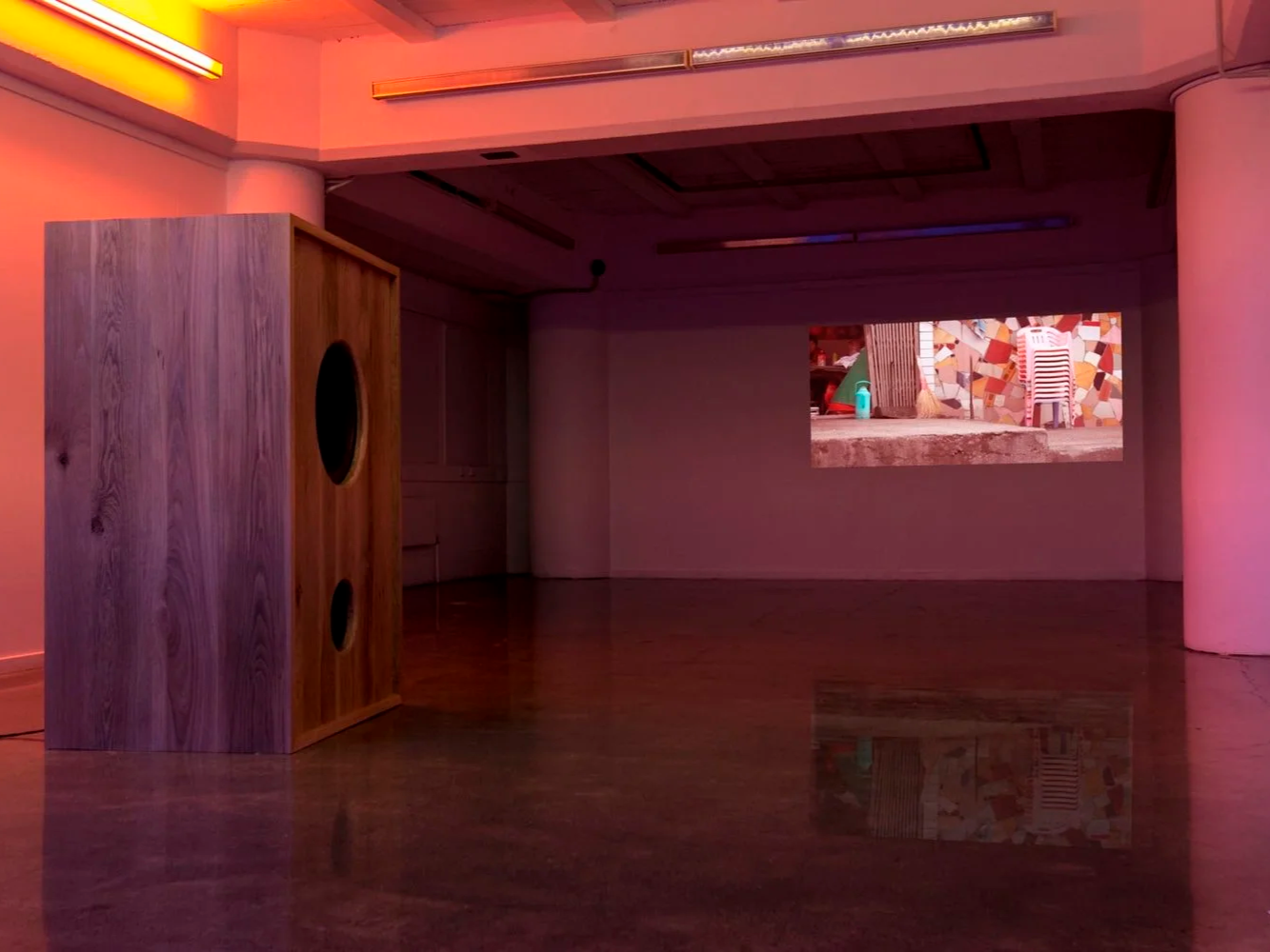 An empty room with pink lighting, a wooden speaker in the foreground, and a video projection on the back wall showing an outdoor scene with a colorful, mosaic-like wall and a plastic chair.