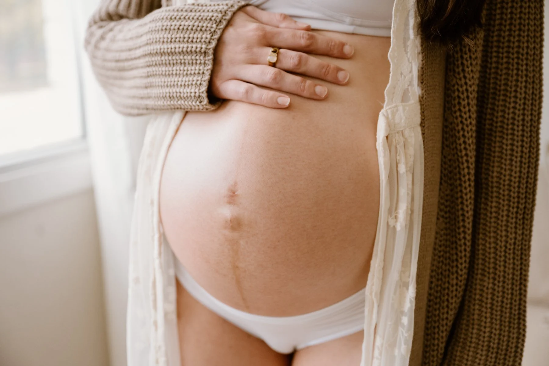 Close-up of a pregnant woman’s belly with her hand resting on it, wearing a ring and a beige cardigan, standing near a white wall and window.