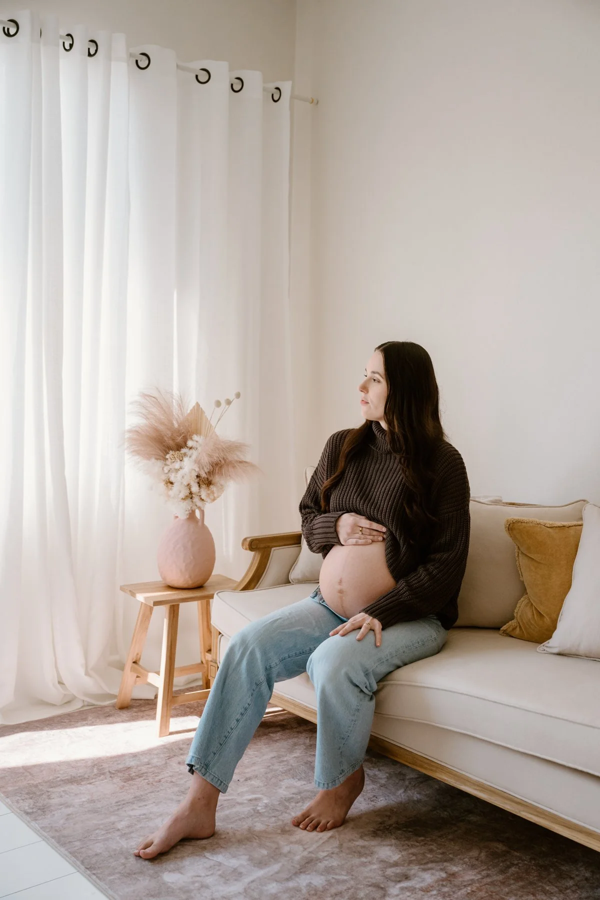 A pregnant woman sitting on a beige sofa in a well-lit living room, holding her belly with one hand and looking to her right. She is wearing a brown sweater and light blue jeans. There is a side table with a pink vase with pampas grass and dried flow
