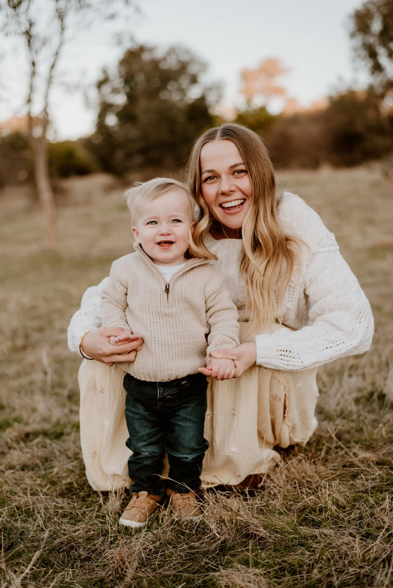 A woman with long, wavy blond hair and a young boy with blond hair smiling outdoors on a grassy field during autumn, wearing beige and neutral-colored sweaters.