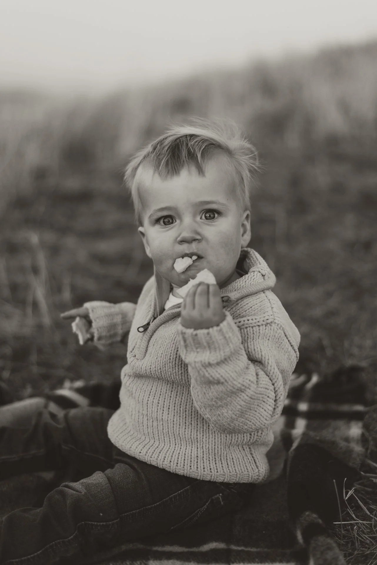 A young child with messy hair and big eyes, wearing a knit sweater and jeans, sitting outdoors on a blanket, eating a snack, with a blurred natural background.
