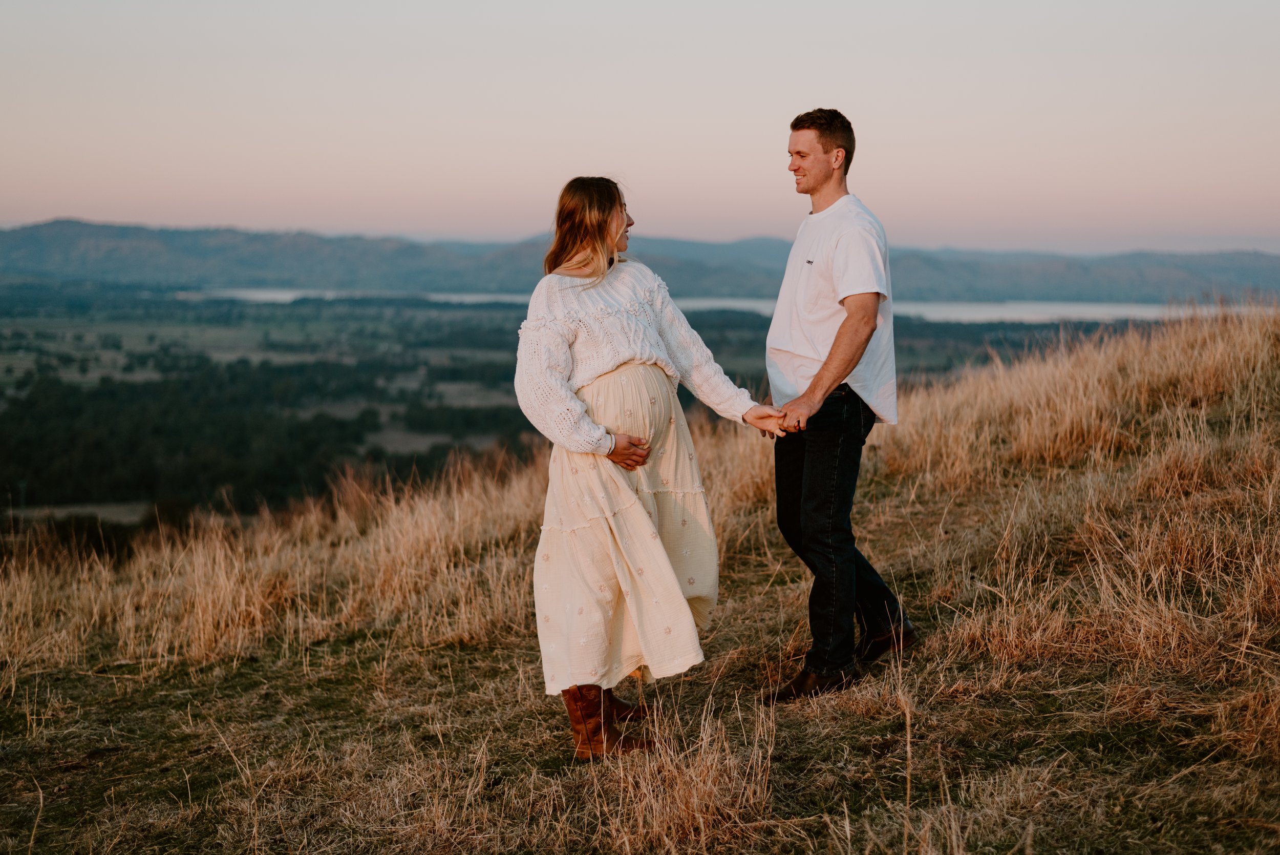 A couple standing outdoors in a grassy field, holding hands and smiling at each other during sunset, with mountains and a lake in the background.