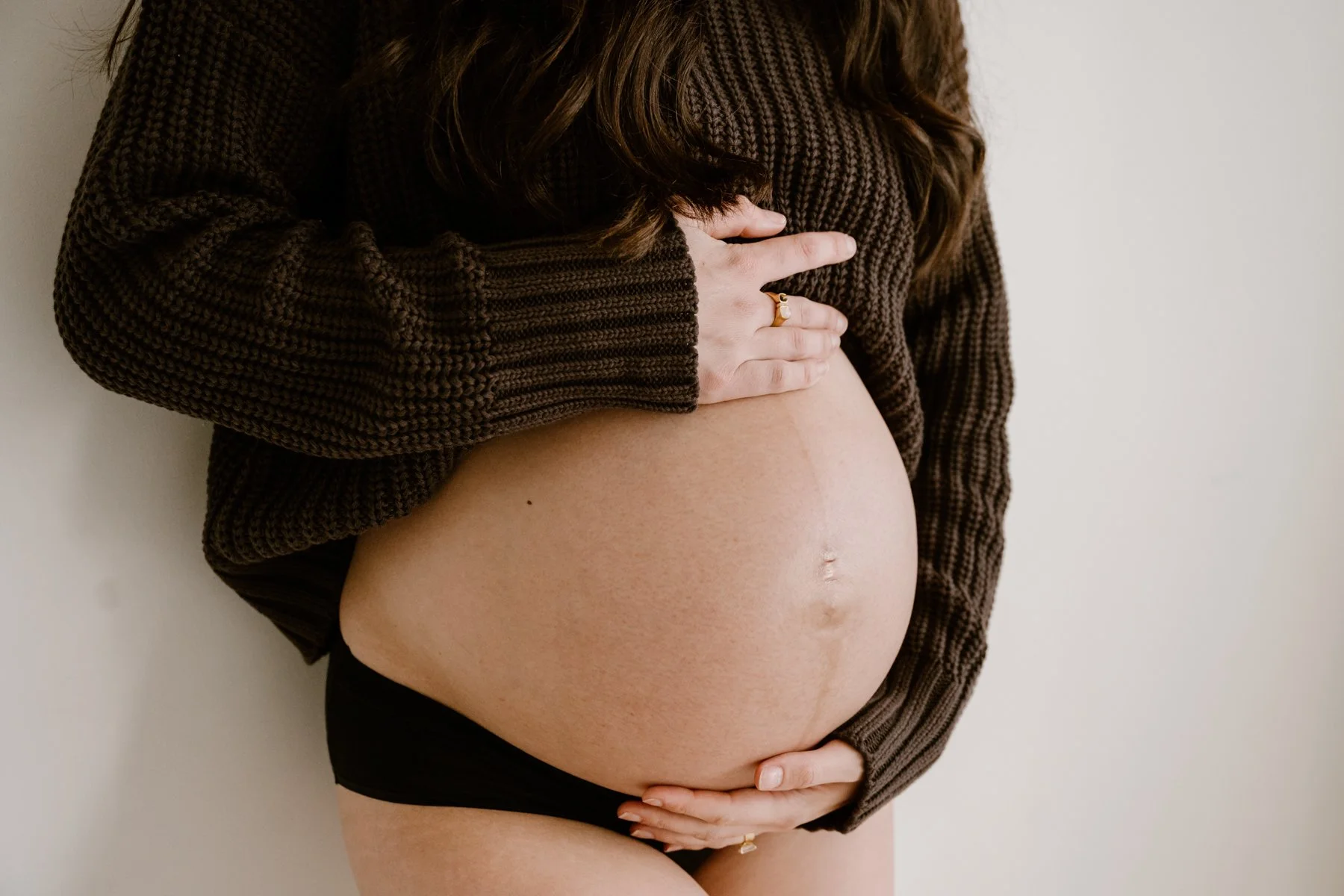 A pregnant woman wearing a brown knitted sweater and black underwear, holding her belly with both hands, against a plain white wall.