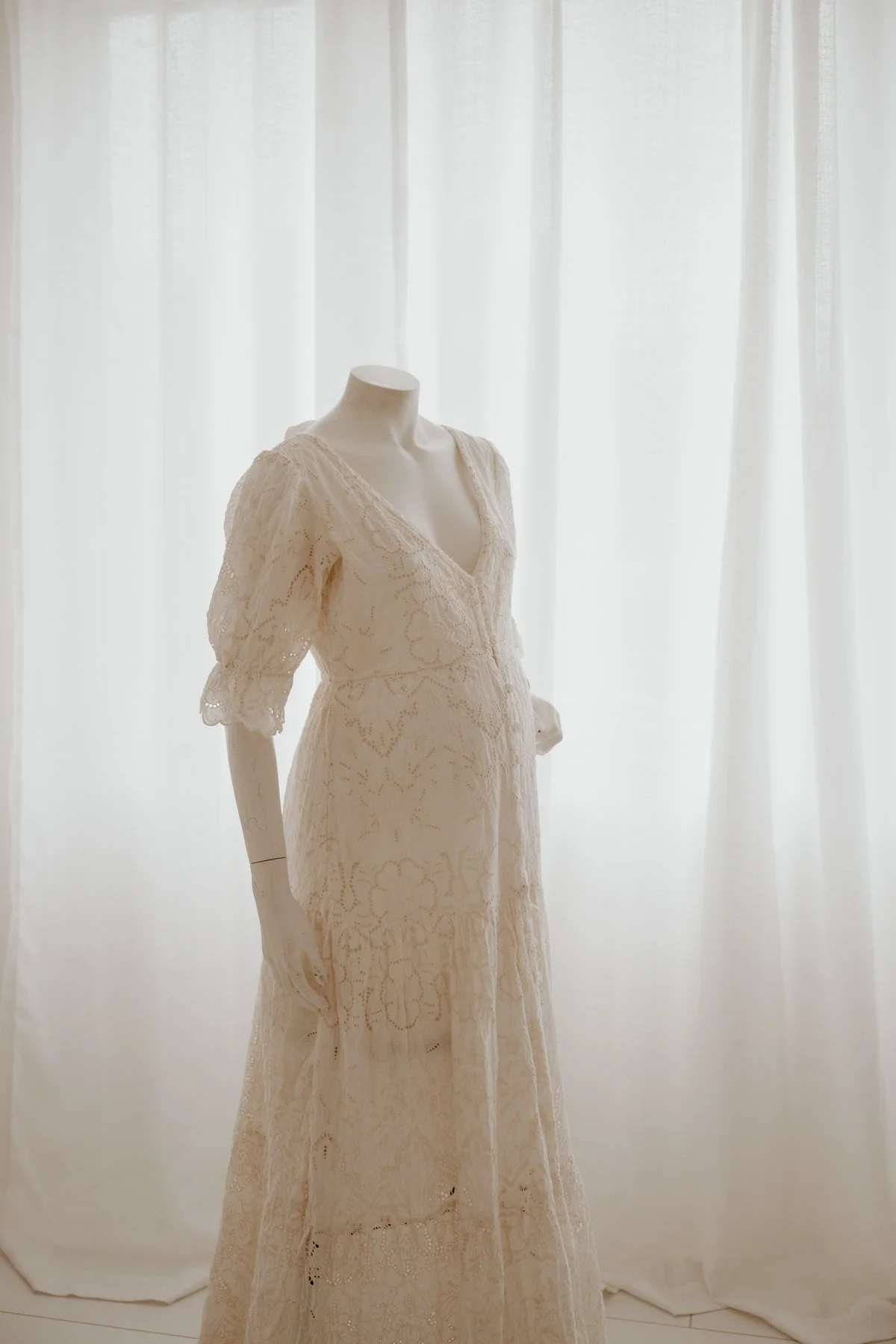 An elegant vintage-style wedding dress with lace details displayed on a mannequin in front of a sheer white curtain.