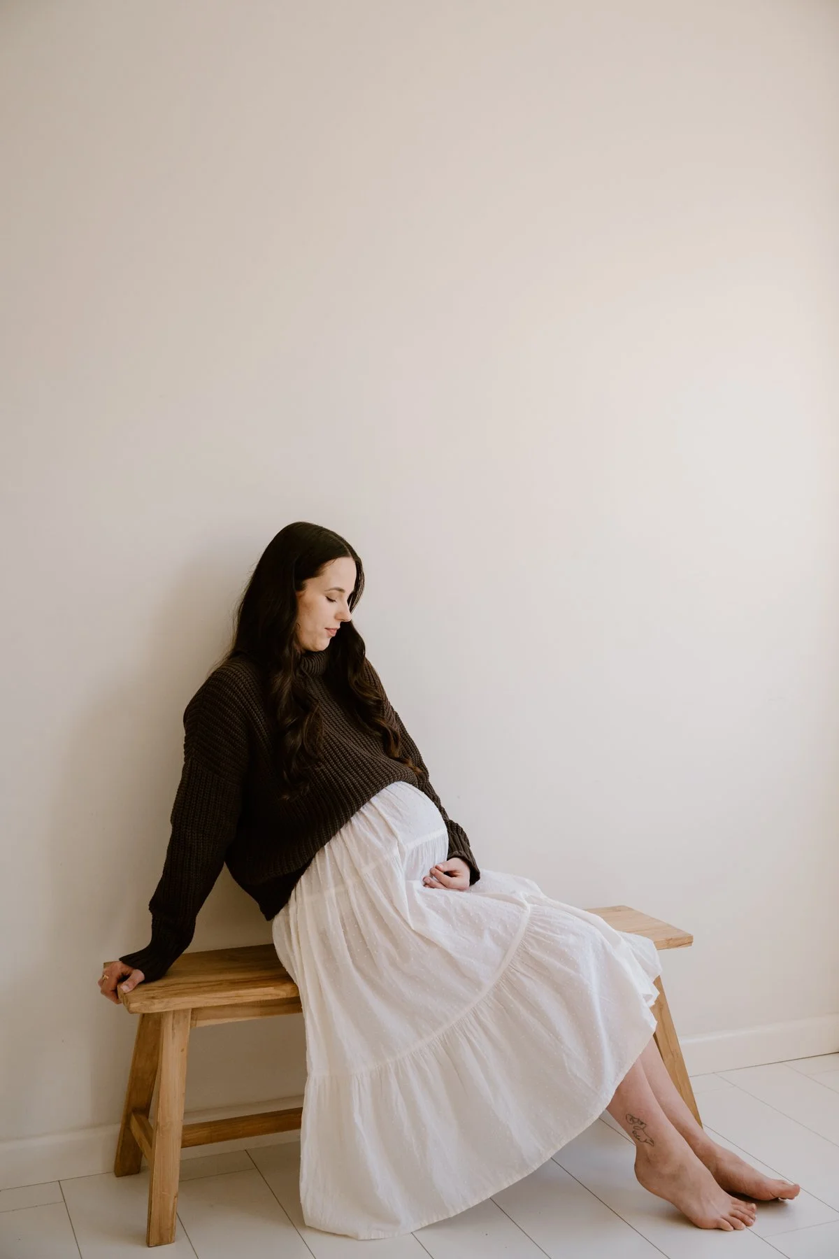 A pregnant woman with long dark hair sitting on a wooden bench against a plain white wall. She is wearing a brown sweater and a flowing white skirt, gazing down at her pregnant belly.