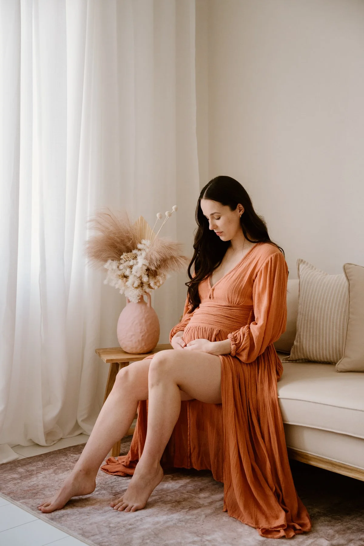 A woman wearing an orange dress sitting on a sofa in a room with natural light. A vase with dried flowers is on a small table beside her, and beige cushions are on the sofa. She is looking down and appears to be pregnant.
