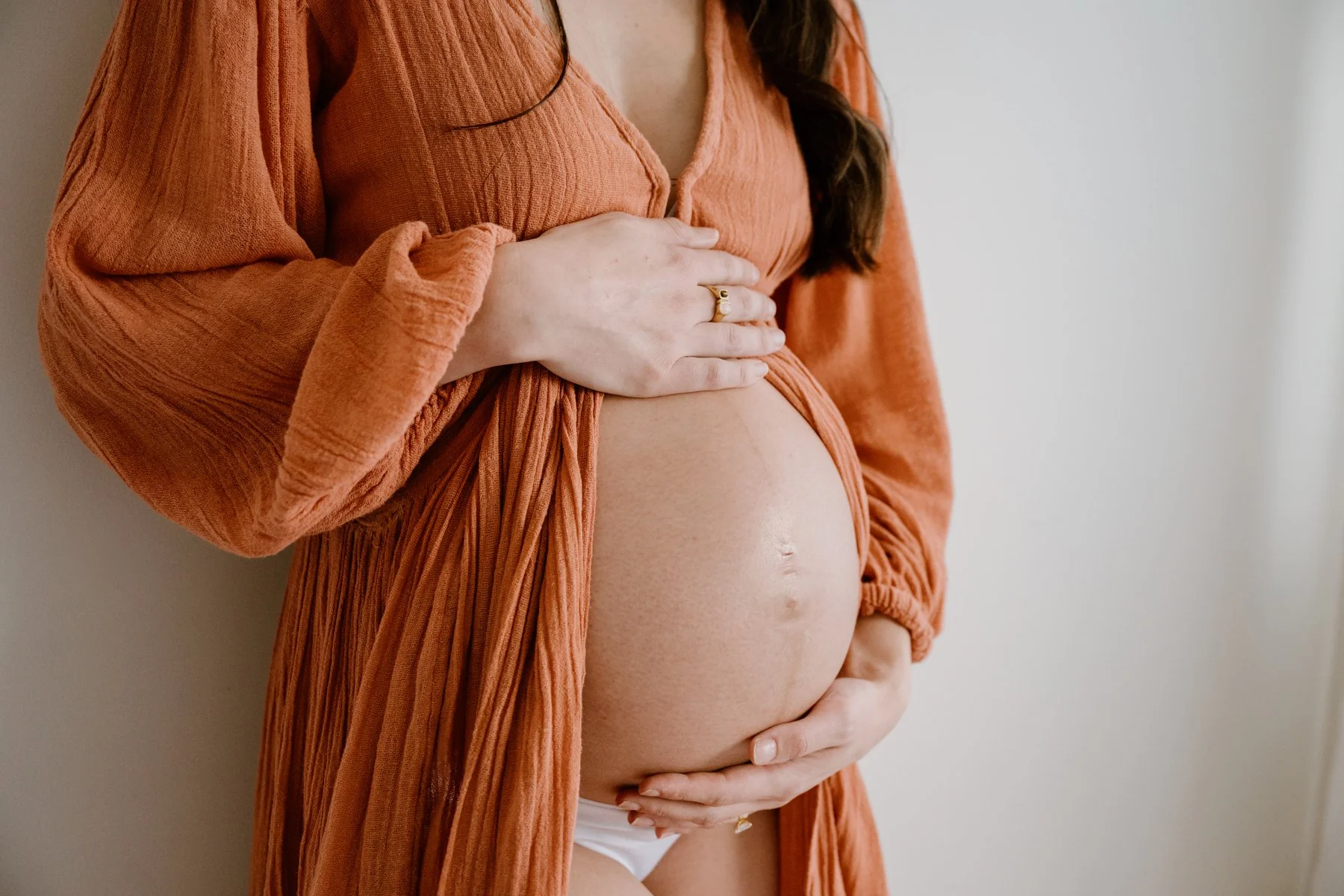 Close-up of a pregnant woman wearing an orange dress, gently touching her baby bump with one hand on her stomach and the other over her chest. The background is plain and neutral.