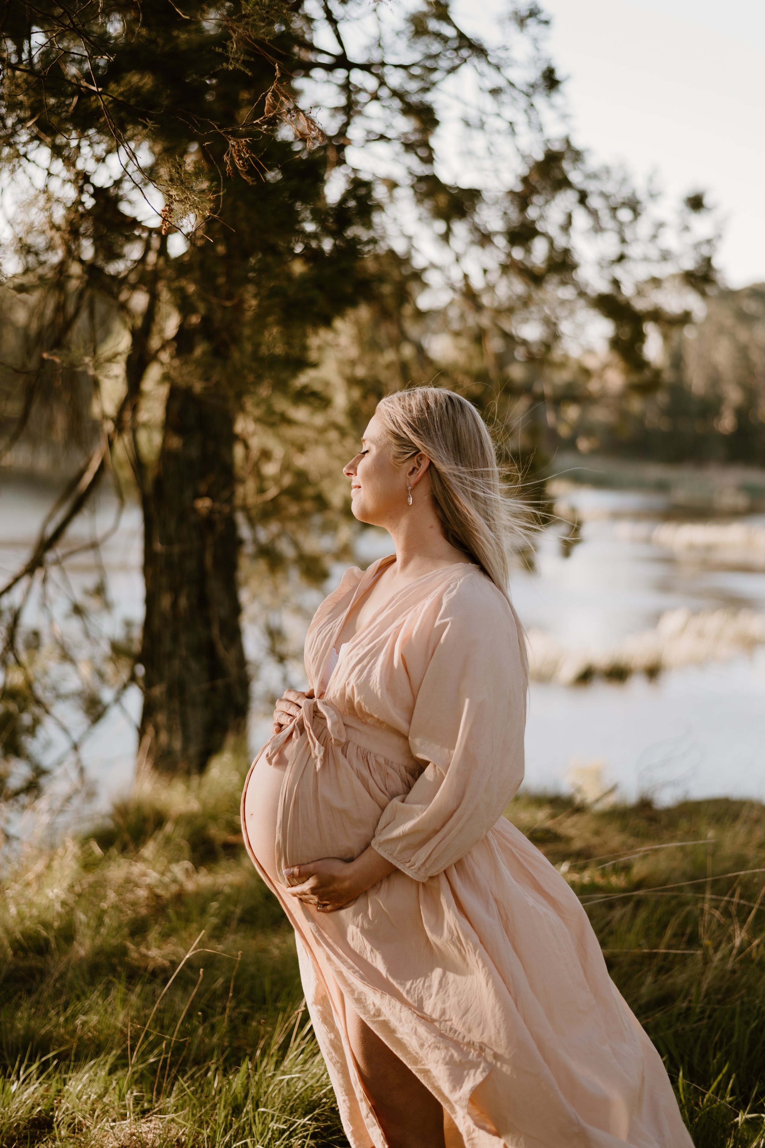 Pregnant woman standing outdoors near Lake Sambell, wearing a light-colored flowing robe, with trees in the background and the sunlight highlighting her profile. Albury-Wodonga maternity photography by Coop & Co Creative