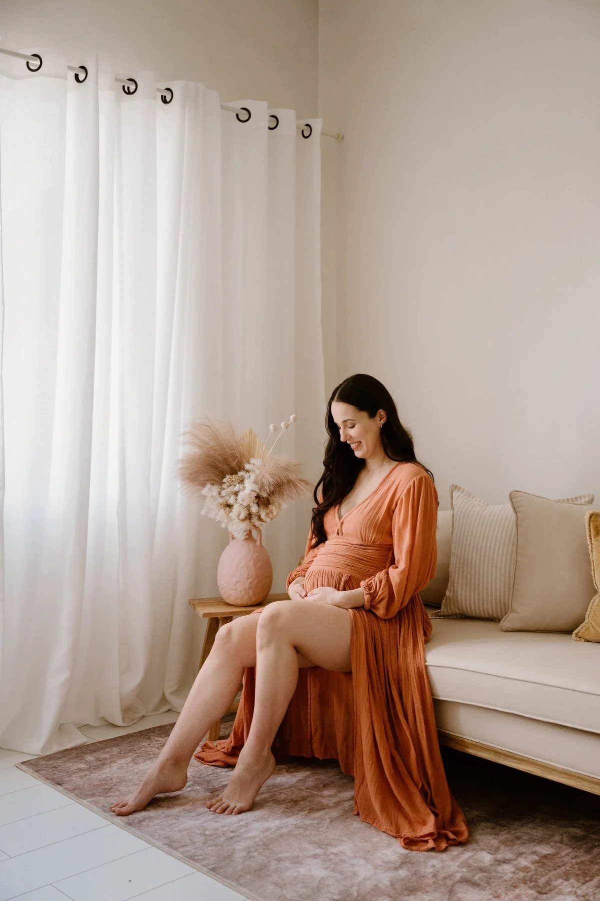 A woman with long dark hair sitting on a beige sofa wearing an orange pleated dress. There is a pink vase with dried flowers on a small wooden table beside her. White curtains hang in the background.