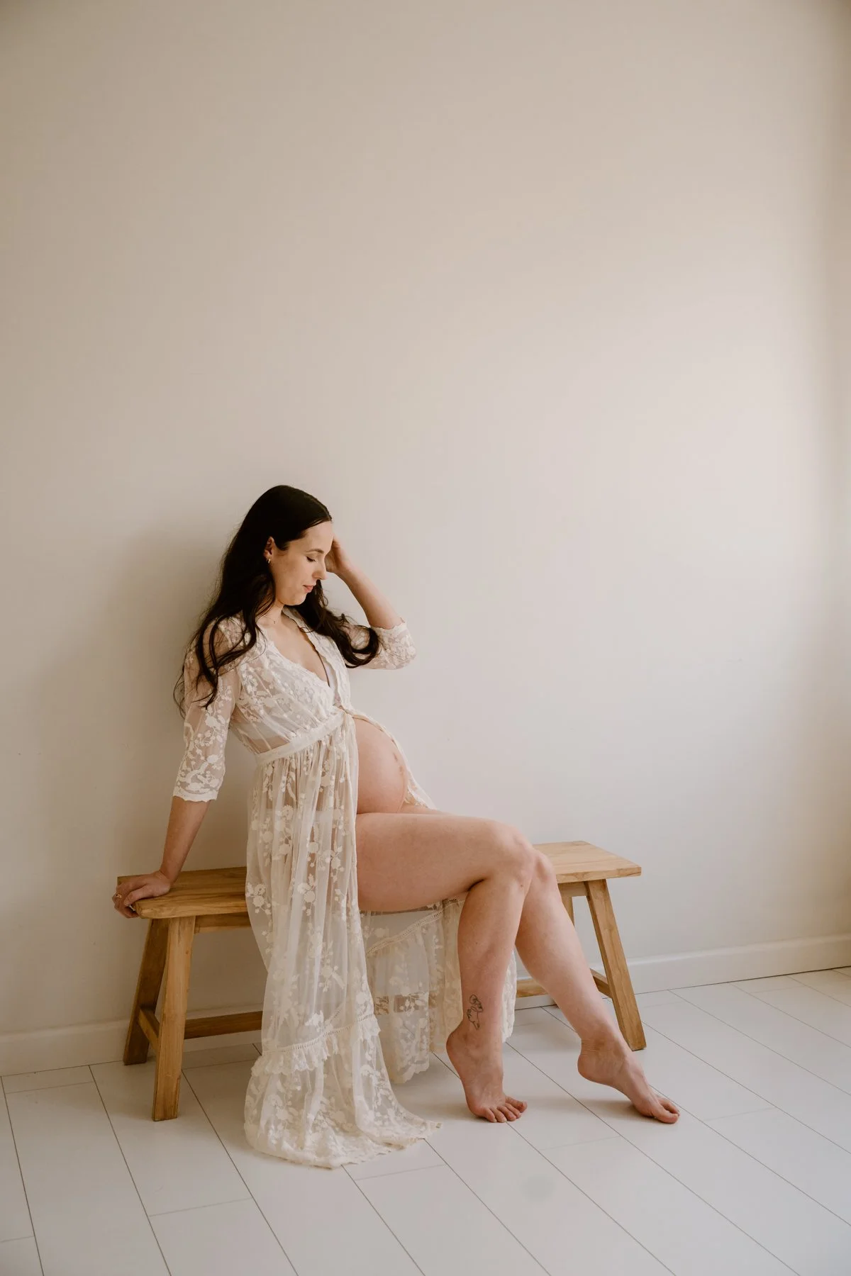 Pregnant woman in lace dress sitting on a wooden bench with a plain white wall behind her, breastfeeding her baby.