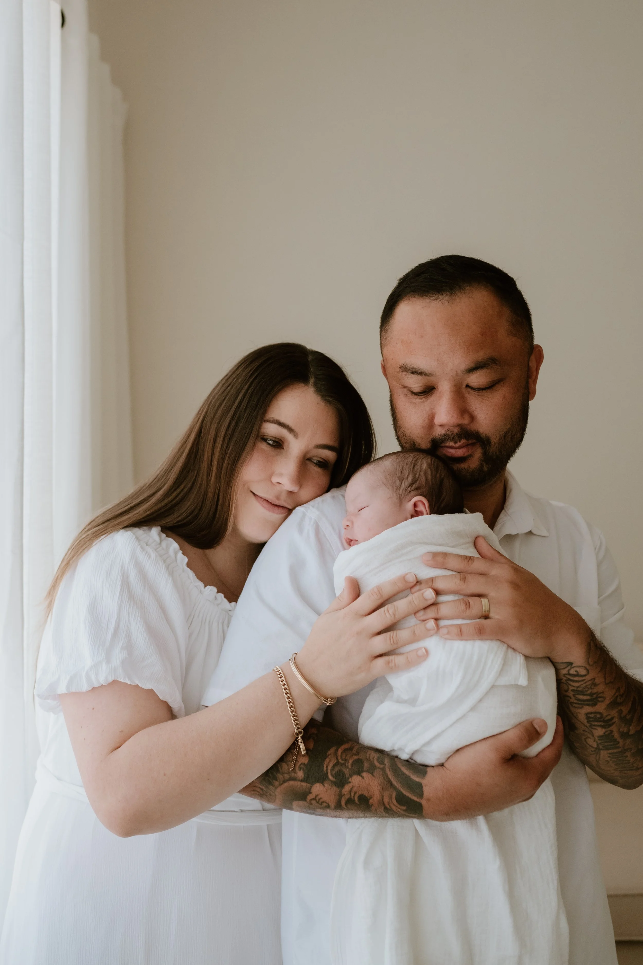 A family of three, a woman, a man, and a baby, close together, with the woman and man holding the baby and looking at it lovingly inside a room with light-colored walls.
