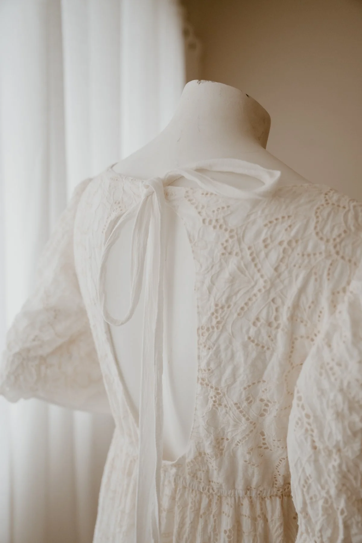 Close-up of a vintage white lace dress on a mannequin, with a bow at the neckline, in front of a curtain.
