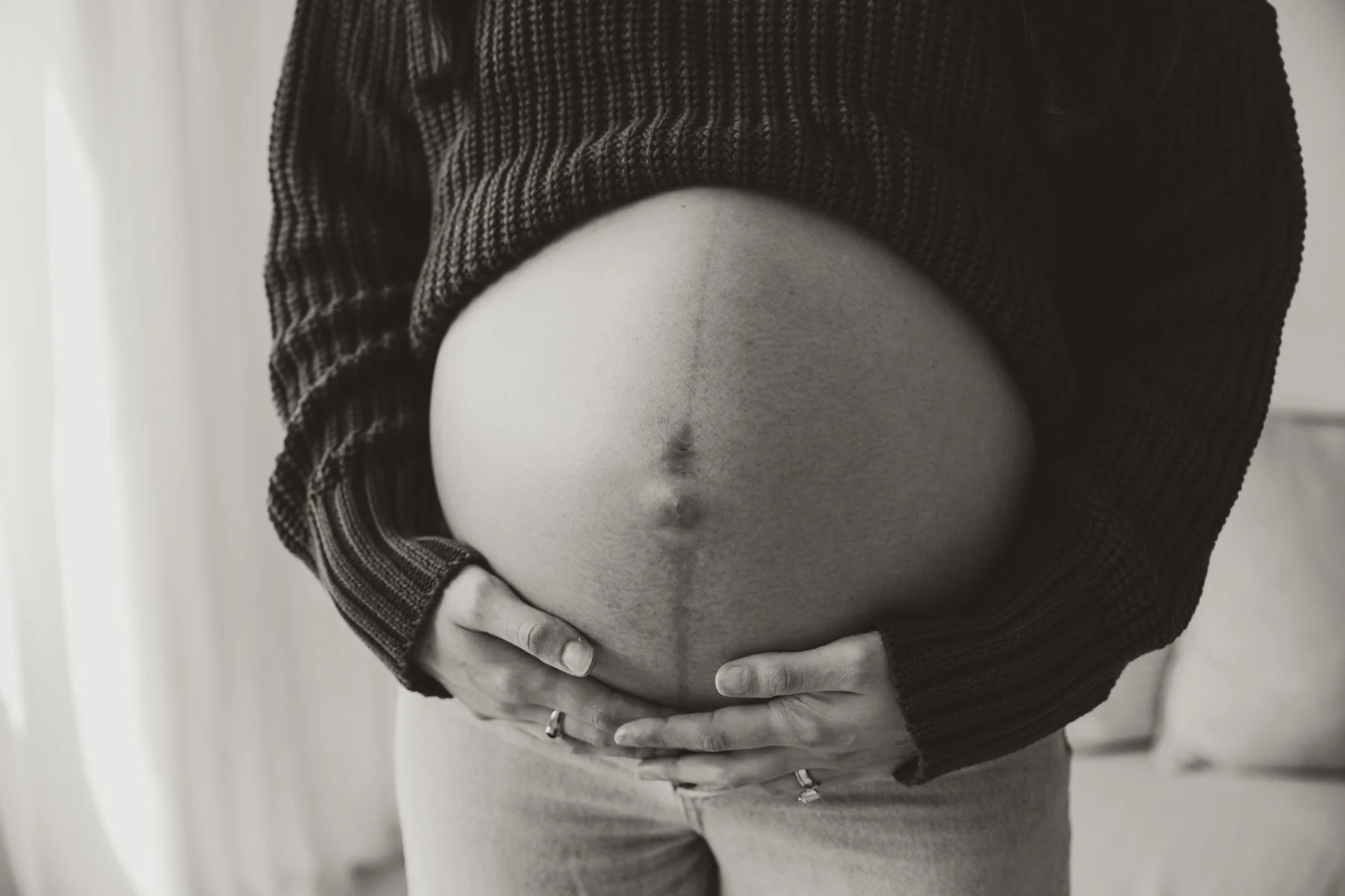 A pregnant woman wearing a dark knurled sweater holding her bare belly with both hands, standing indoors.