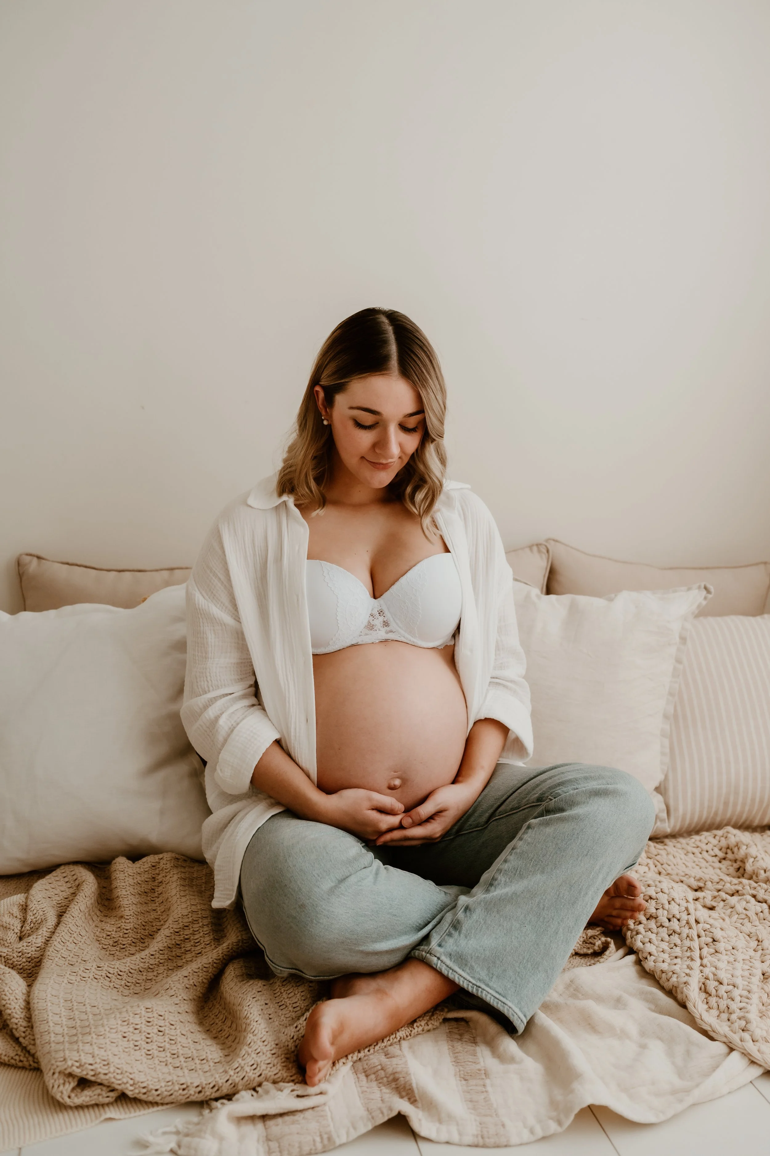 A pregnant woman sitting cross-legged on a bed, gently holding her belly, wearing a white bra, an open white shirt, and gray pants, with pillows and knitted blankets around her.