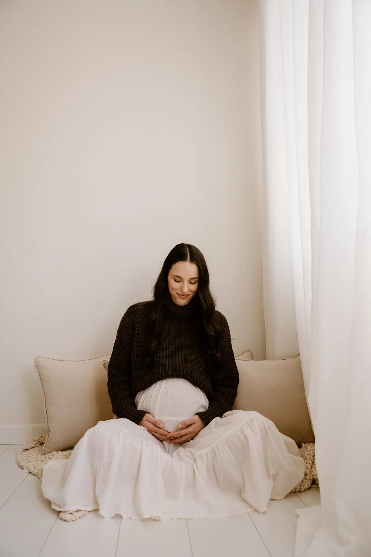 A pregnant woman sitting on a beige couch, smiling and gently holding her belly with her hands, in a softly lit room with white curtains.