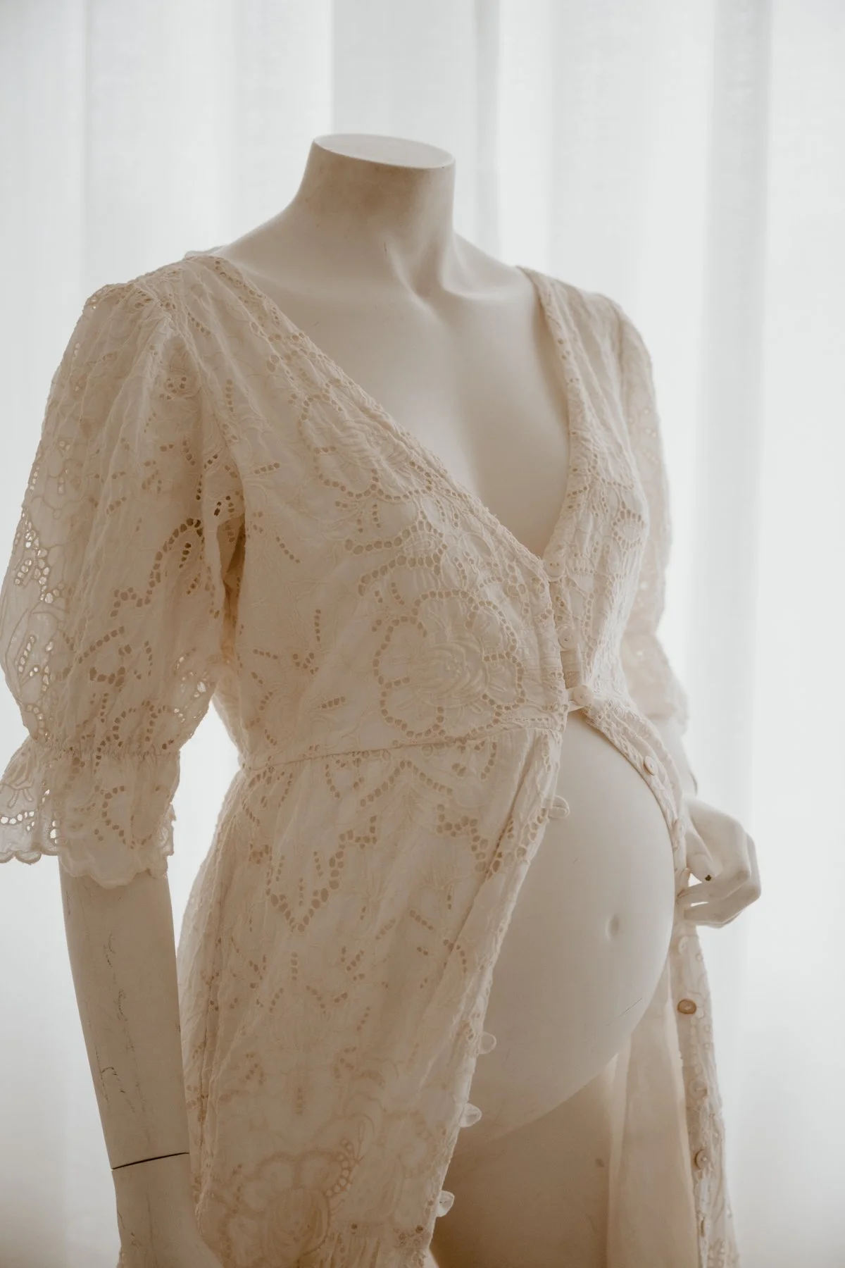 Cream lace maternity dress on a mannequin with a baby bump, in front of a white curtain.