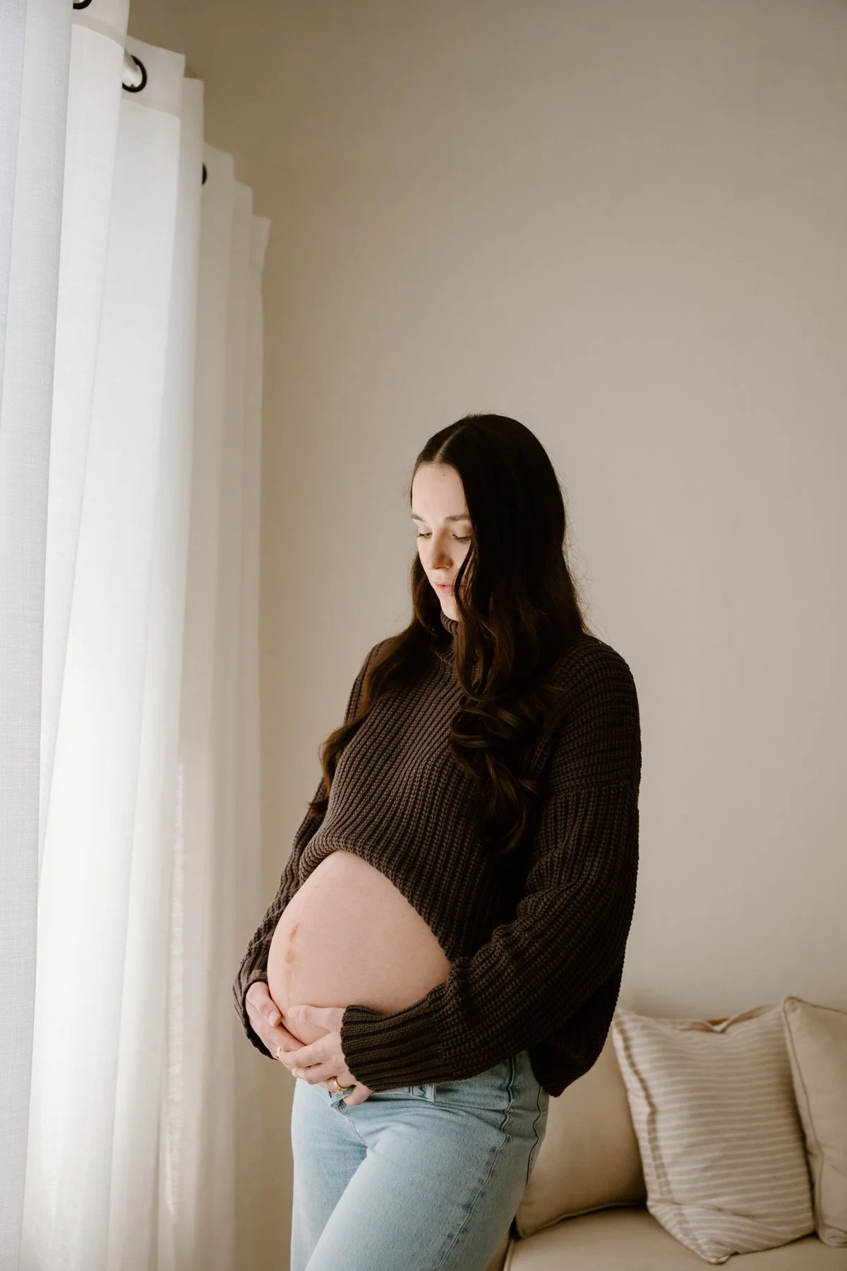 A pregnant woman with long dark hair stands indoors near a window with white curtains, gently holding her pregnant belly and looking down thoughtfully.