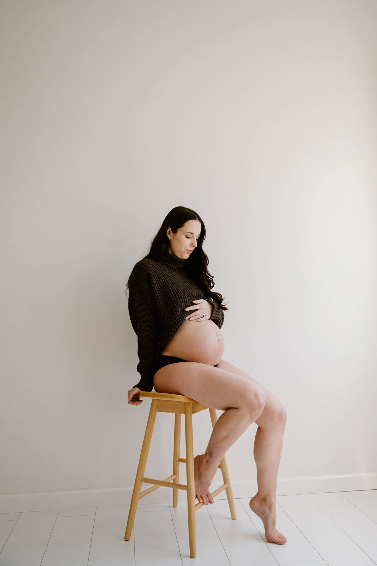 A pregnant woman with long dark hair, wearing a black sweater and black underwear, sitting on a wooden stool. She is cradling her baby bump with her right hand and has her left hand resting on the stool, with a serene expression on her face, against 