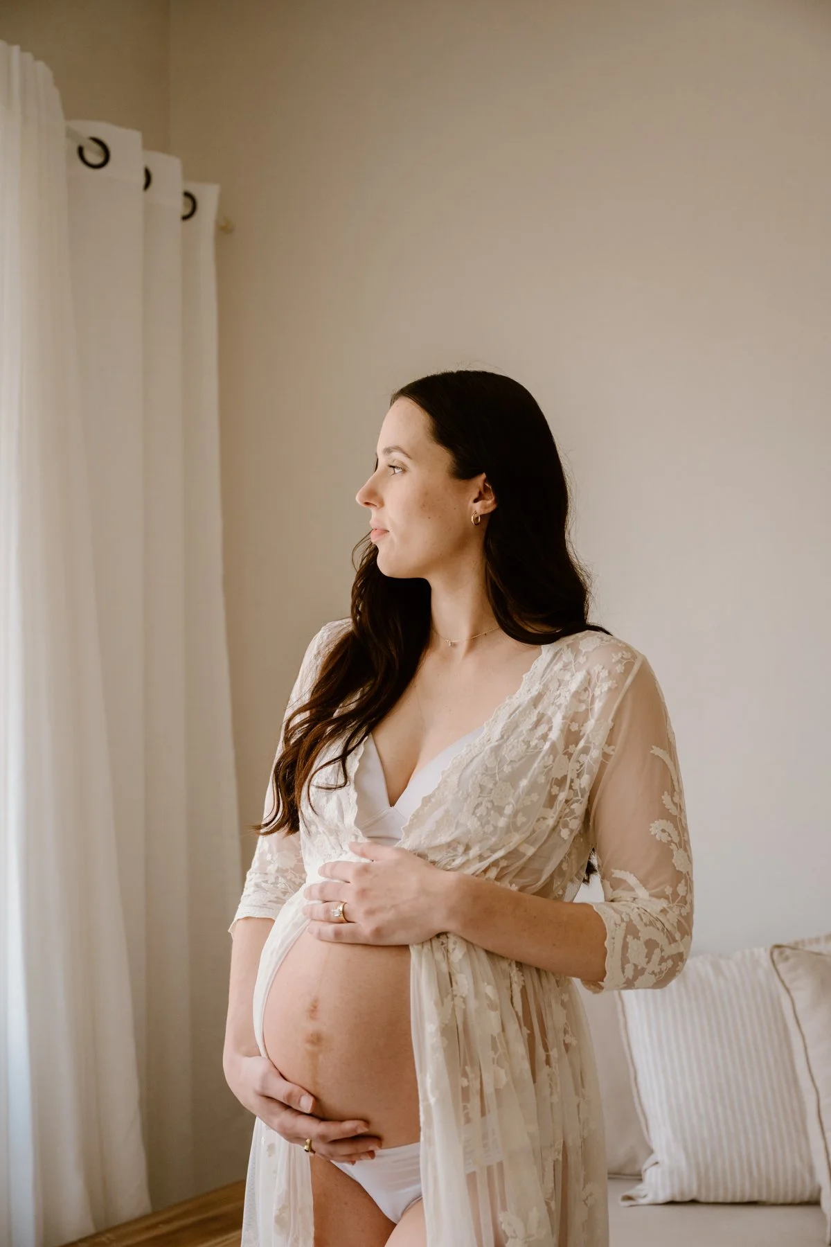 A pregnant woman with long dark hair in a cream lace robe holding her belly, standing near a window with white curtains, in a softly lit bedroom.