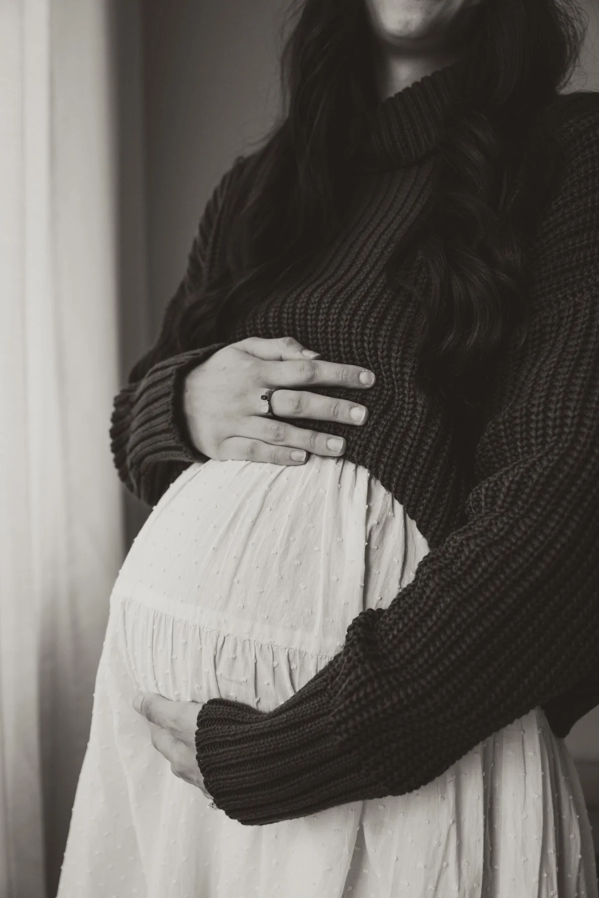 A pregnant woman wearing a dark knitted sweater and a light-colored, textured skirt, cradling her baby bump with both hands.