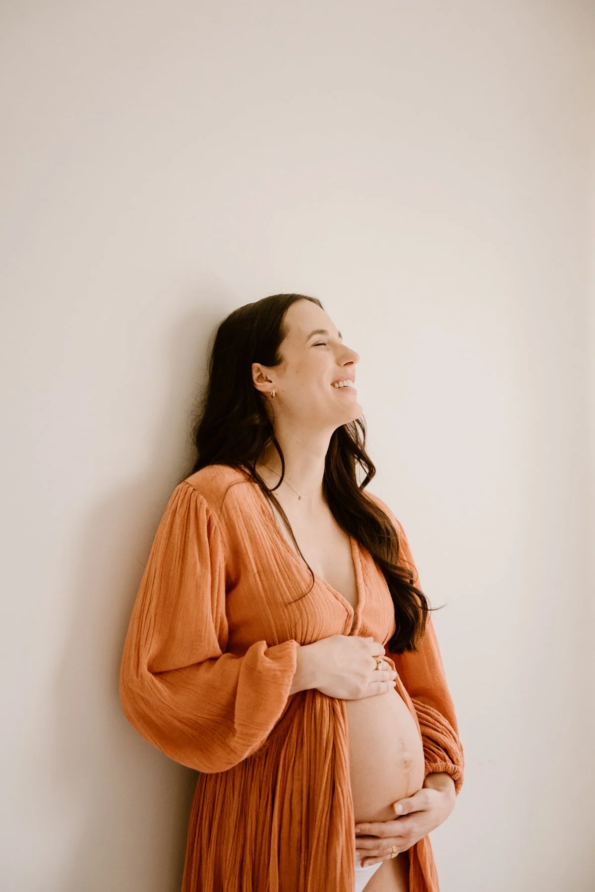 A pregnant woman with long dark hair, wearing a rust-colored dress, smiling with her eyes closed, cradling her baby bump with both hands, standing against a plain light-colored wall.