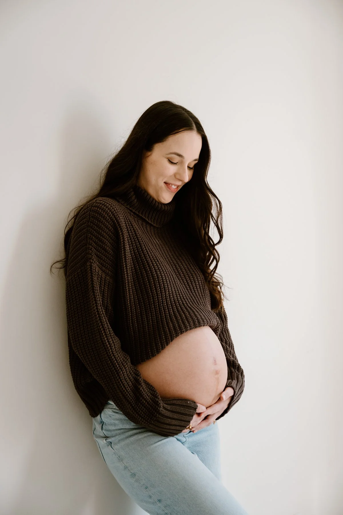 Pregnant woman with long dark hair wearing a brown sweater and light blue jeans, standing against a plain white wall, smiling and holding her belly.