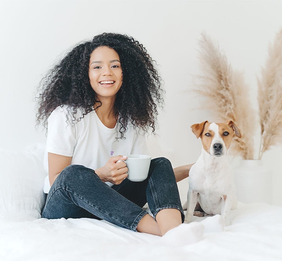 A beautiful young wife who loves to wear her husband's shirt is sitting on the bed with her dog. She's in a super comfortable and soft white tee - her husband's graphic undershirt. The saying on the shirt is "Be the Change".