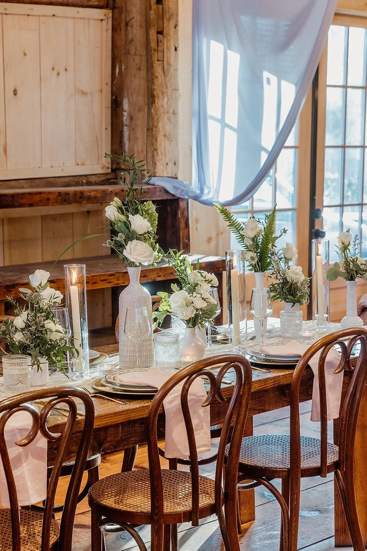 Rustic dining table decorated with white floral centerpieces, candles, and glassware, set beside wooden walls and a window with flowing white curtains.