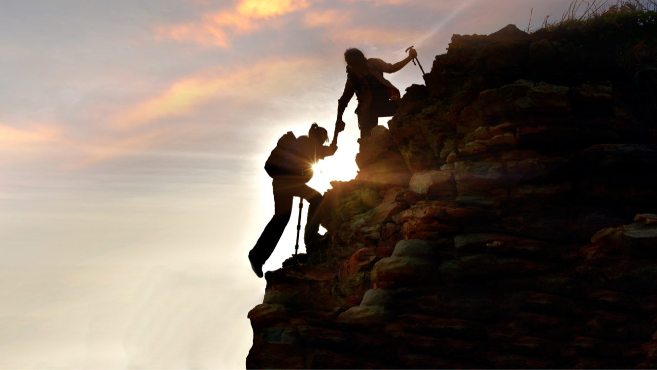 Two climbers helping each other ascend a rocky cliff at sunset, one reaching down to assist the other.