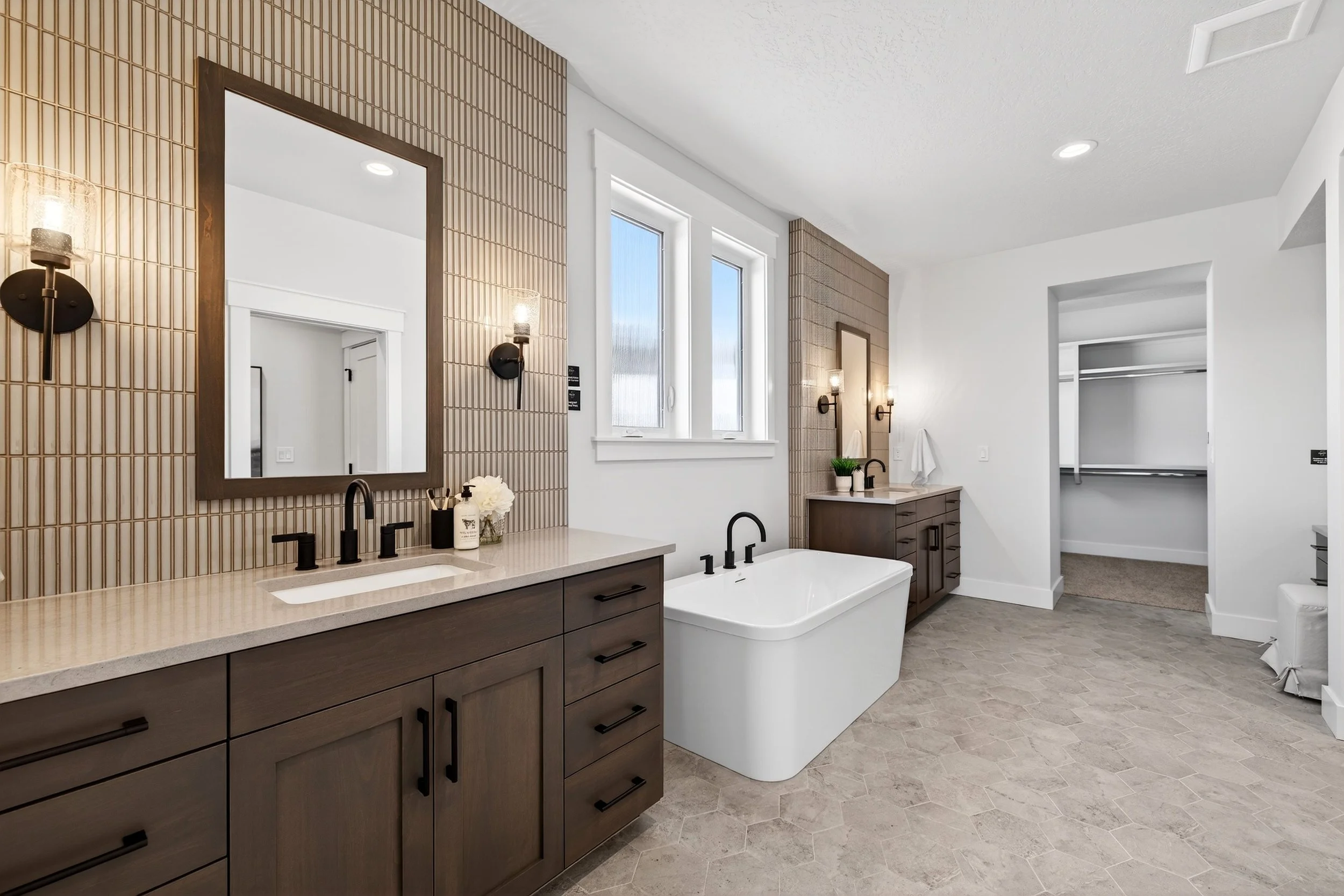 Modern bathroom with wooden cabinetry, a white bathtub with black fixtures, a large mirror, wall sconces, and a walk-in closet.