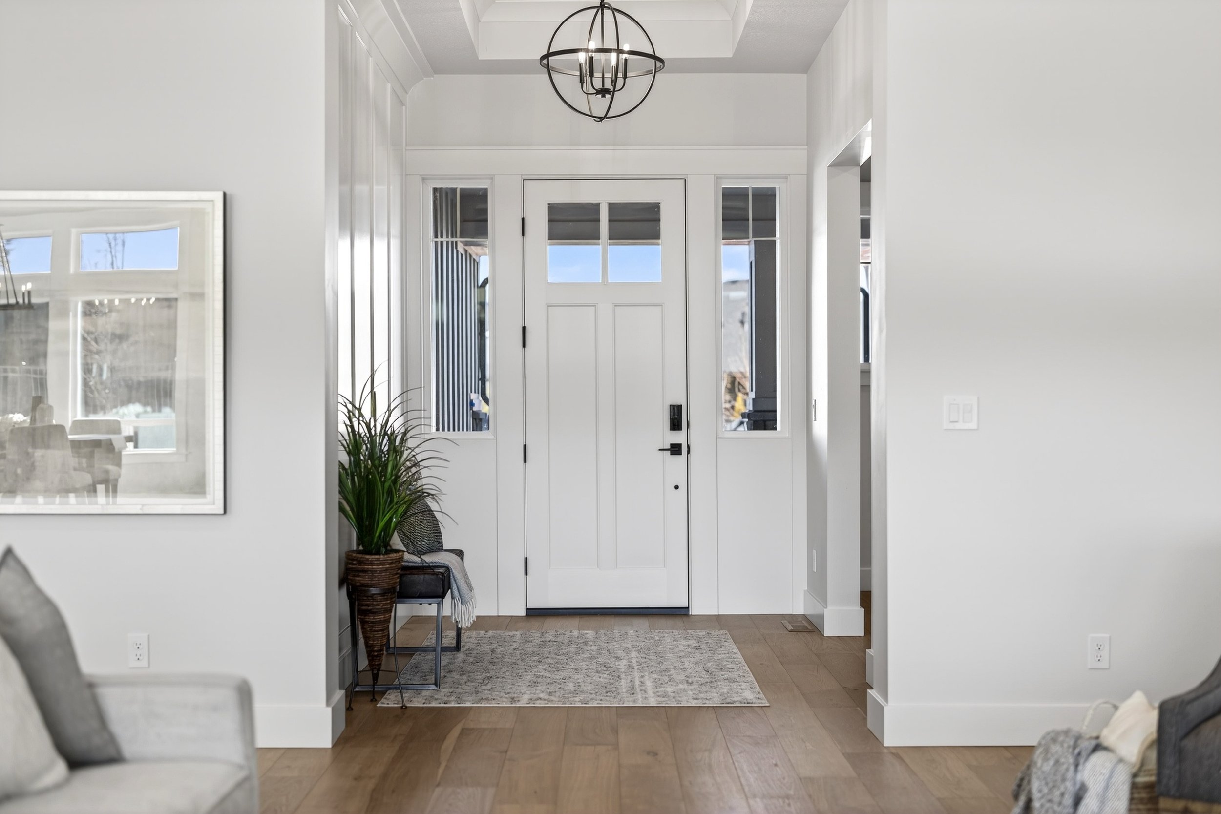 Modern white front door with glass panels and black hardware, flanked by tall sidelights, in a bright entryway with wood flooring, a potted plant, a chair, and a ceiling light fixture.
