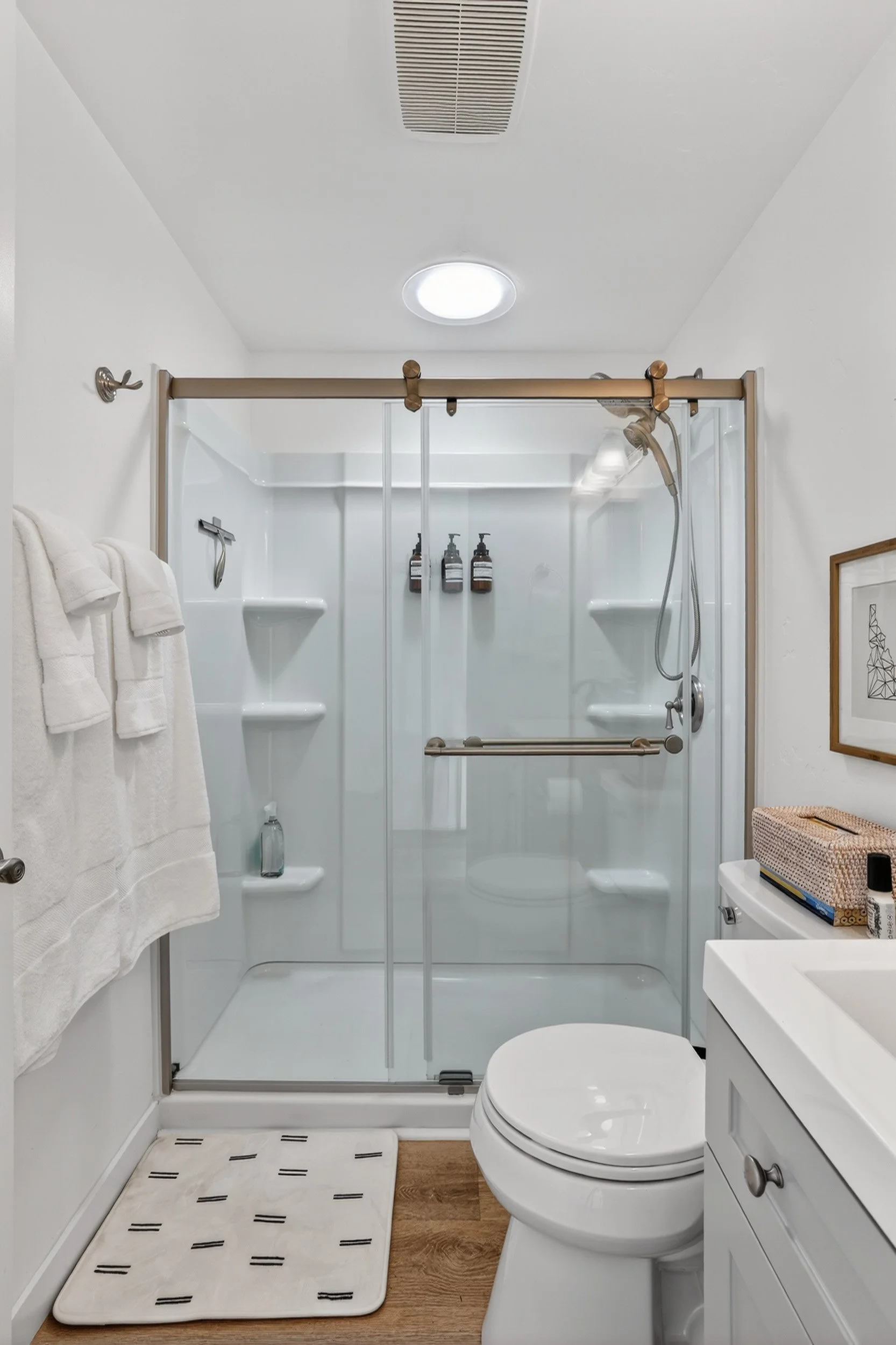 A clean, modern bathroom with a shower glass enclosure, white walls, a white toilet, a white sink, and wooden accents on the slide door, towel rack, and floor.